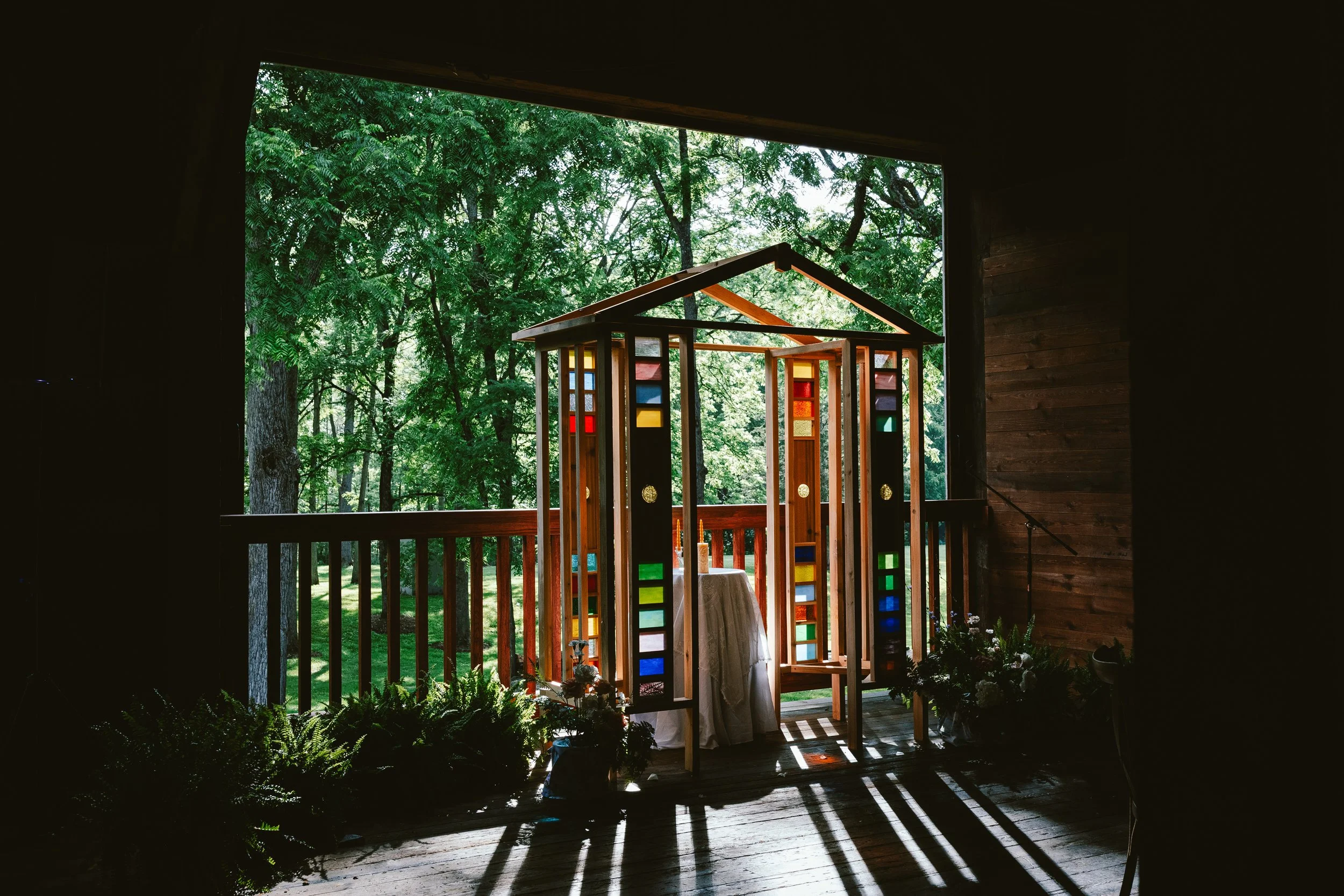 A colorful stained glass altar on a wooden porch with sunlight casting shadows, surrounded by greenery.