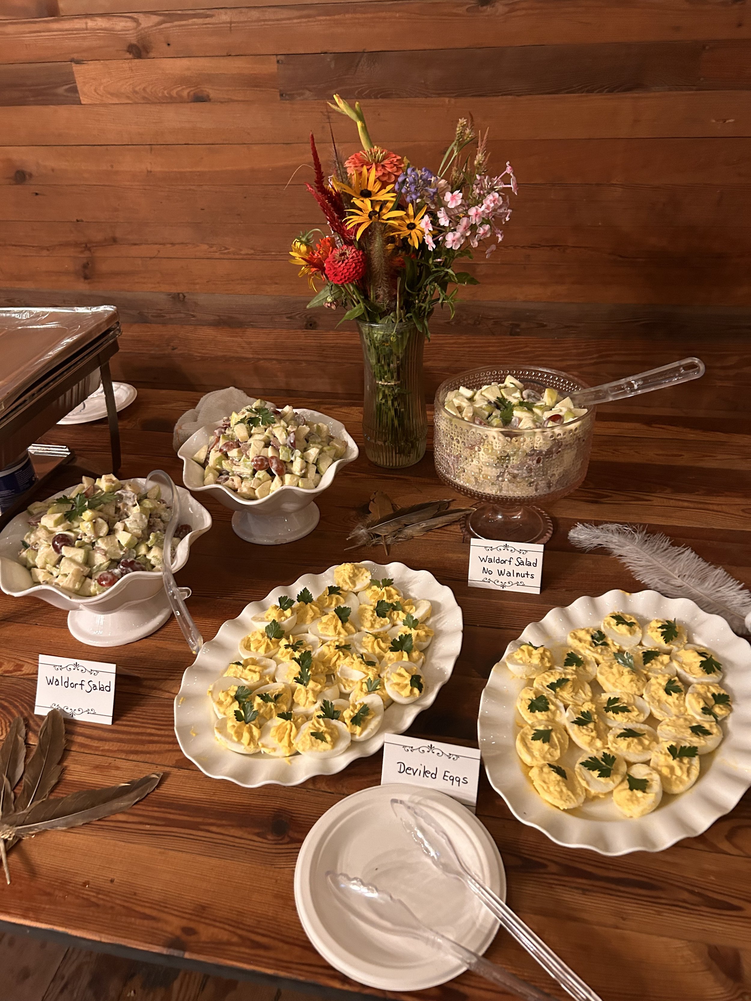 A wooden table with plates of Waldorf salad and deviled eggs, accompanied by a vase of flowers. The salads are labeled: 'Waldorf Salad' and 'Waldorf Salad No Walnuts.' The deviled eggs are garnished with parsley.