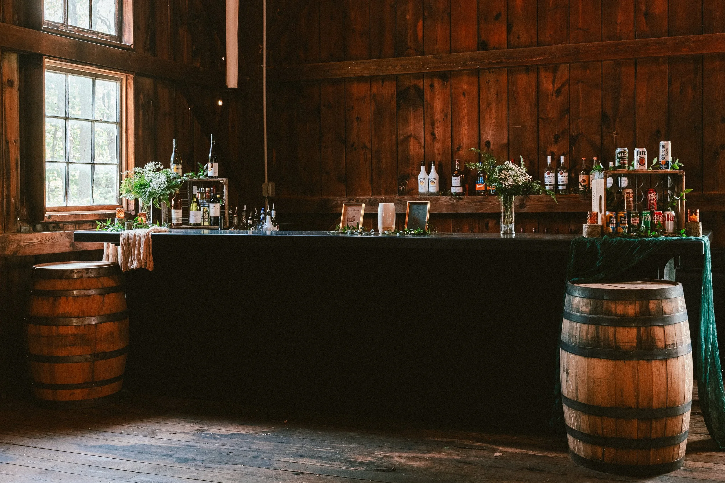 A rustic wooden bar inside a cabin with alcohol bottles, flowers, and bar accessories, flanked by two wooden barrels, and illuminated by natural light from windows.