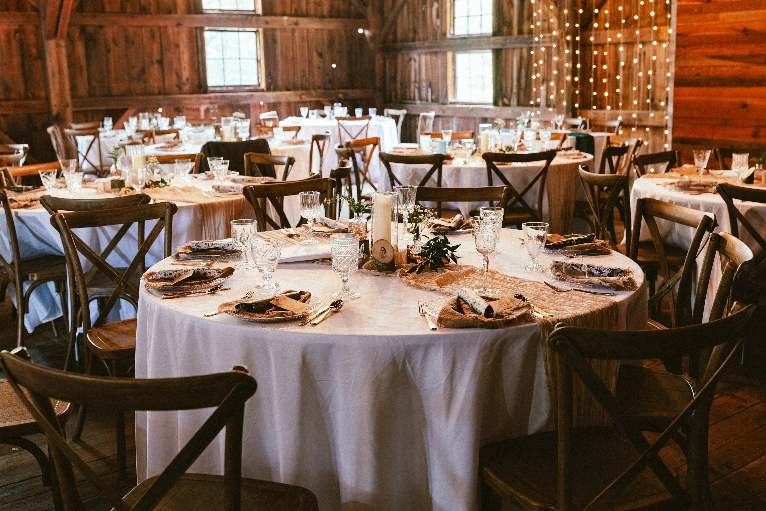 Round tables set with white tablecloths, glassware, silverware, paper napkins, and centerpieces in a rustic wooden venue decorated with string lights.