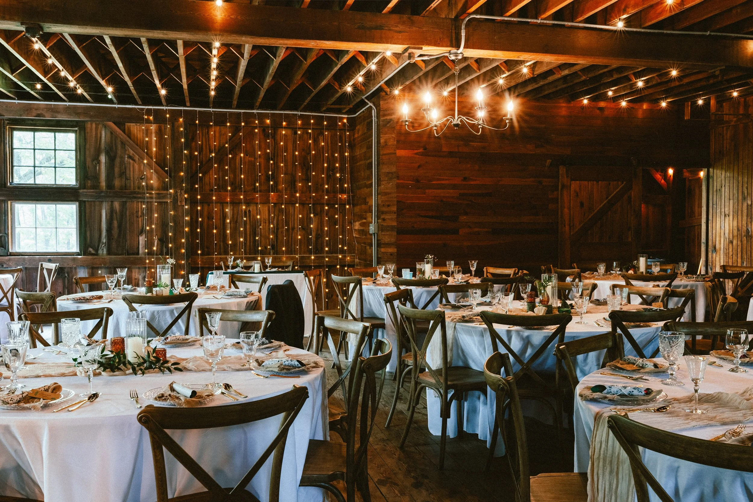 A rustic banquet hall decorated for a celebration with round tables covered in white tablecloths, set with glassware, silverware, and napkins. The wooden walls and ceiling are accented with string lights and a chandelier, creating a warm and inviting