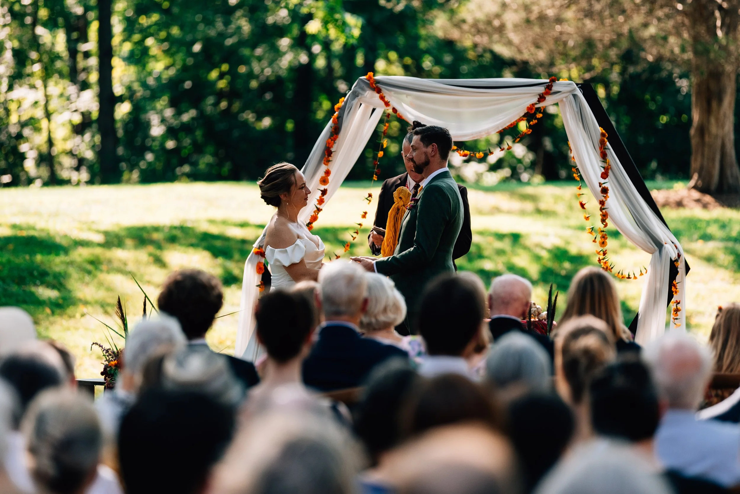 A couple gets married outdoors during a wedding ceremony, standing under a decorated archway with orange flowers, with guests seated watching in the background.