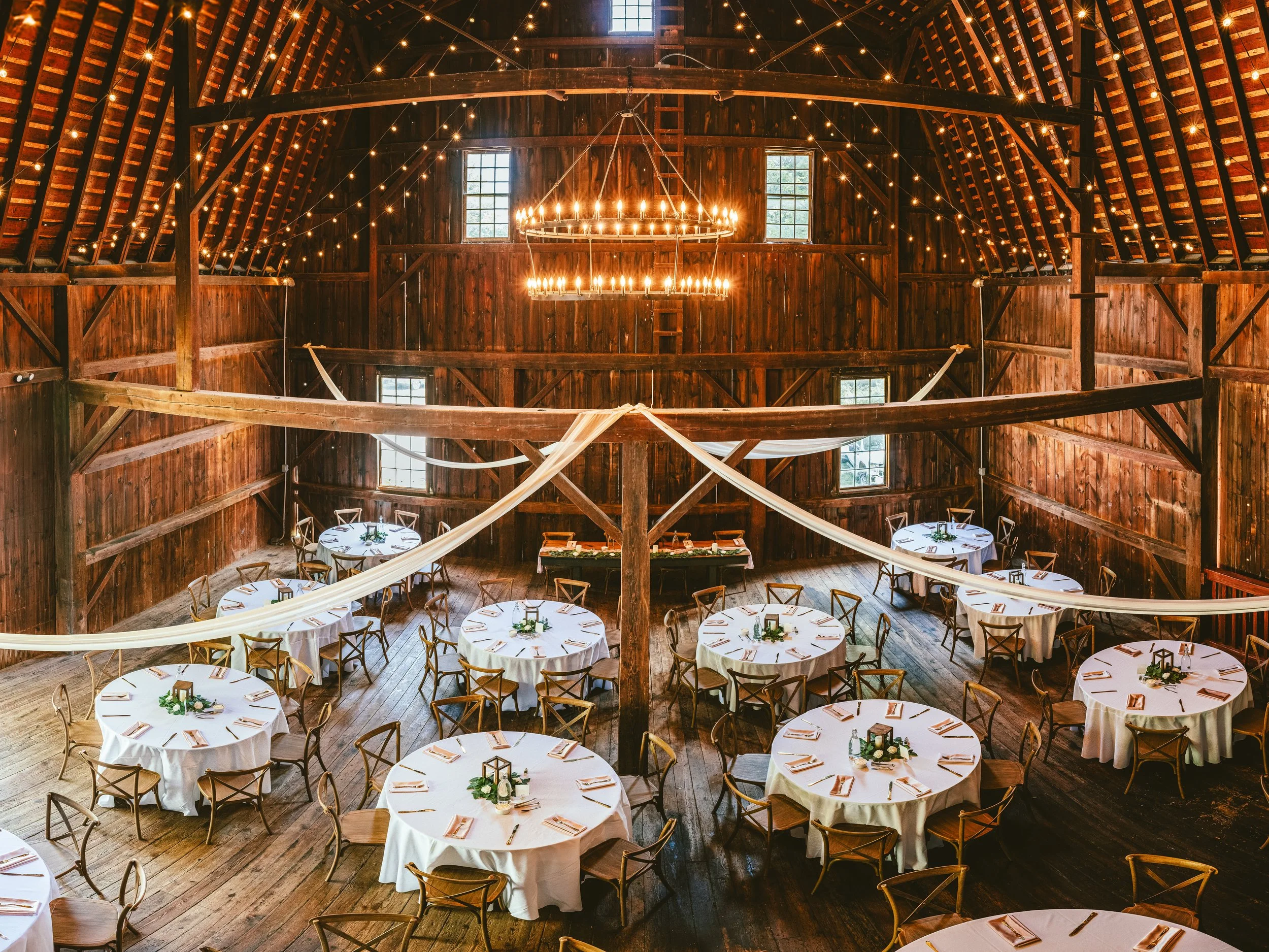 Wedding reception setup inside a rustic barn with wooden walls, round tables with white tablecloths, centerpieces, and wooden chairs. Dim lighting with string lights and a large chandelier hanging from the ceiling.