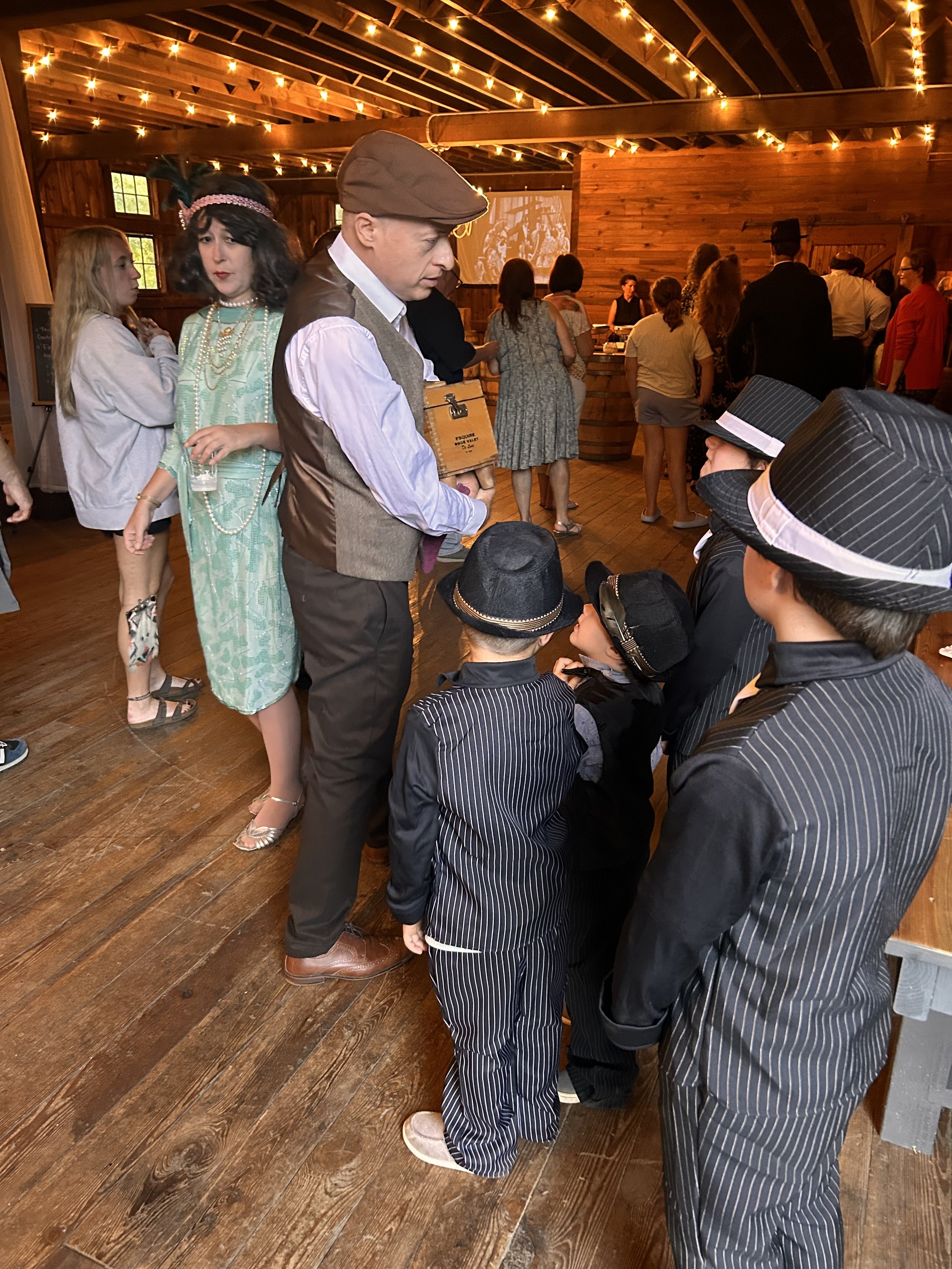 A group of people dressed in 1920s-style clothing inside a rustic venue decorated with string lights. A man in a cap and vest interacts with several children wearing pinstripe suits and hats. Other adults and children in period attire are gathered ar