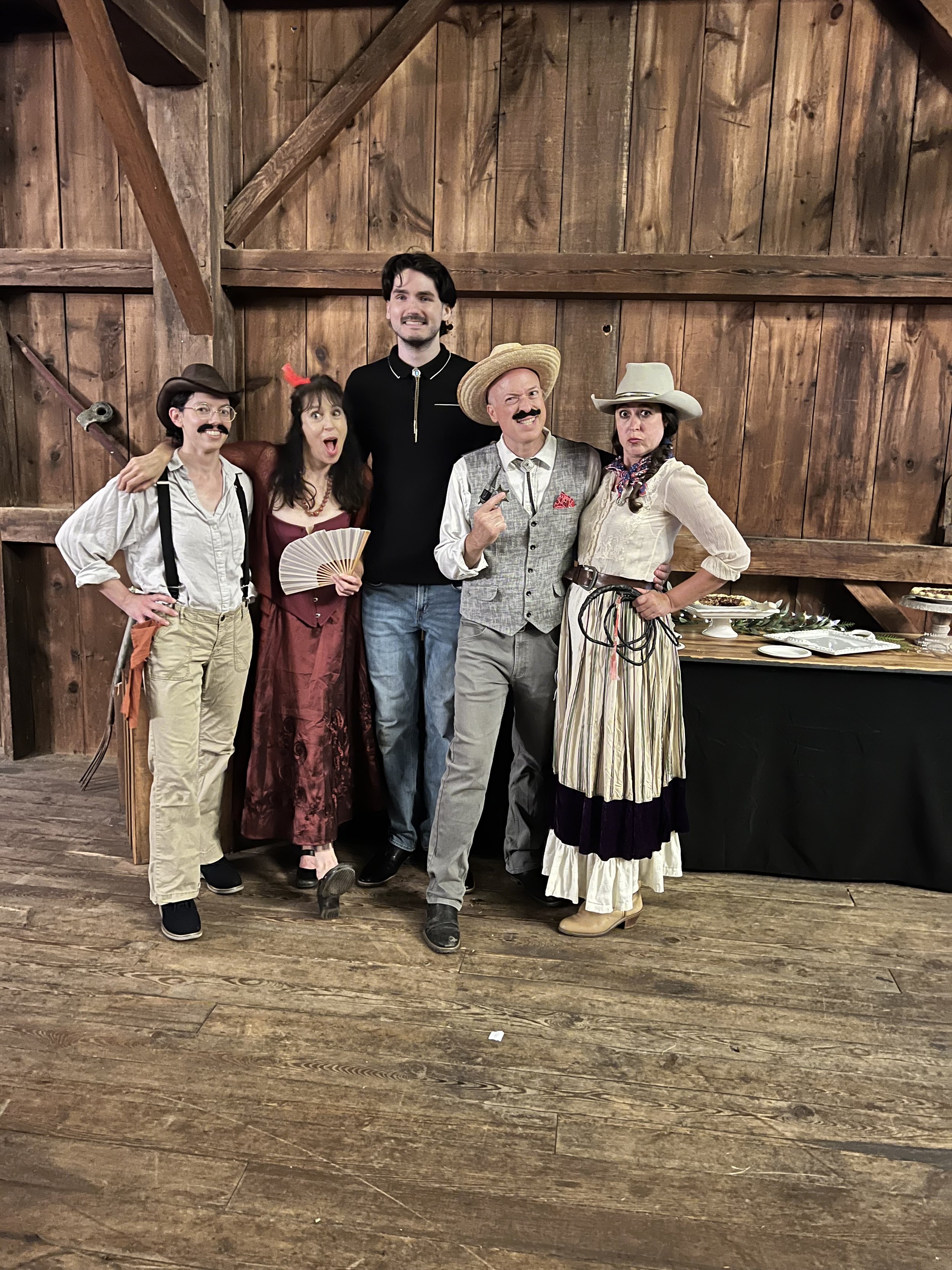 A group of five people in Western-style costumes pose in front of a wooden backdrop. One person wears suspenders and a hat, another holds a fan in a red dress, the central person wears a modern black shirt, and the others are in cowboy attire with ha