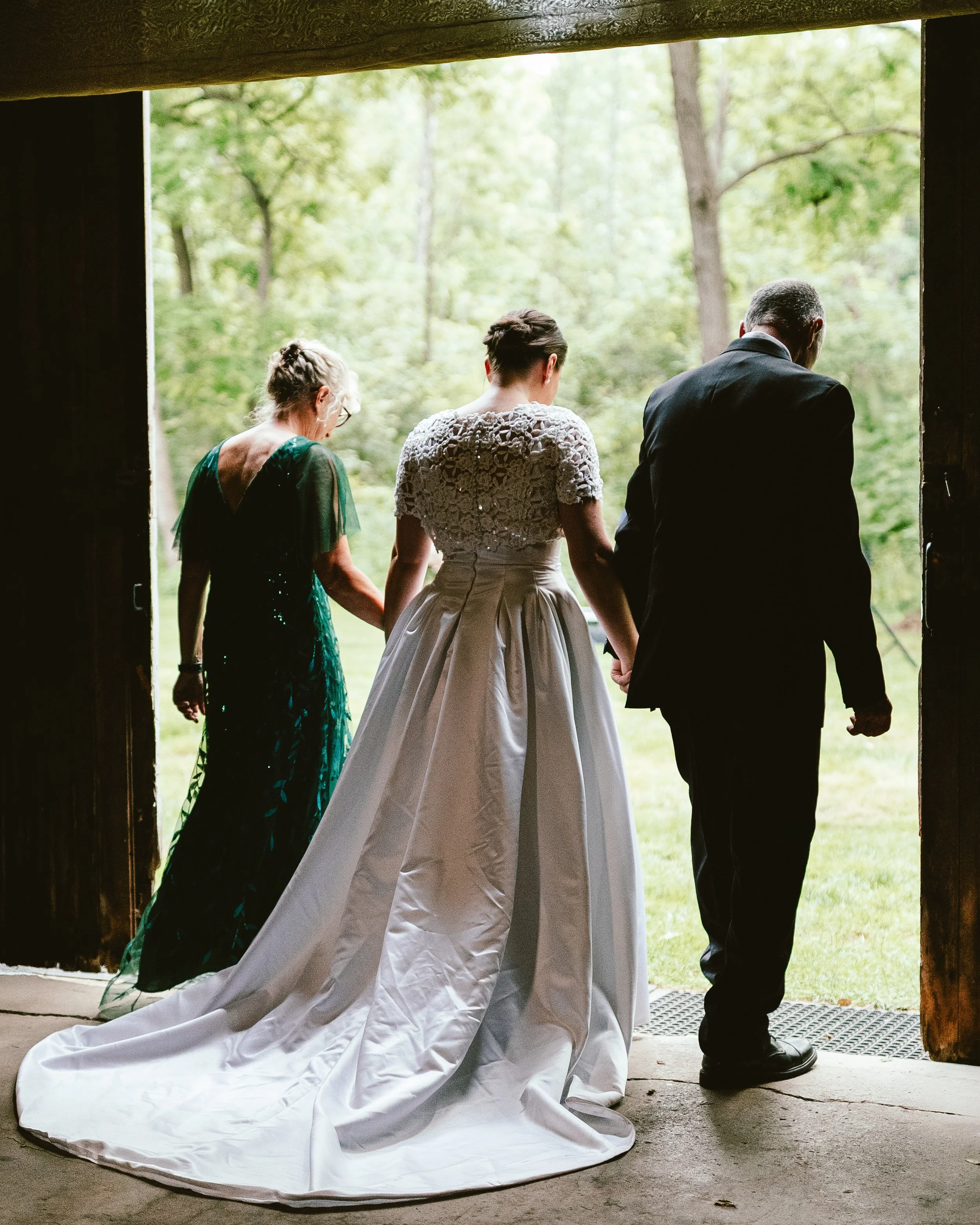 A bride in a white wedding gown holding hands with an older man in a black tuxedo, with a woman in a dark green dress standing beside them, all walking out of a rustic wooden structure into a green outdoor space.