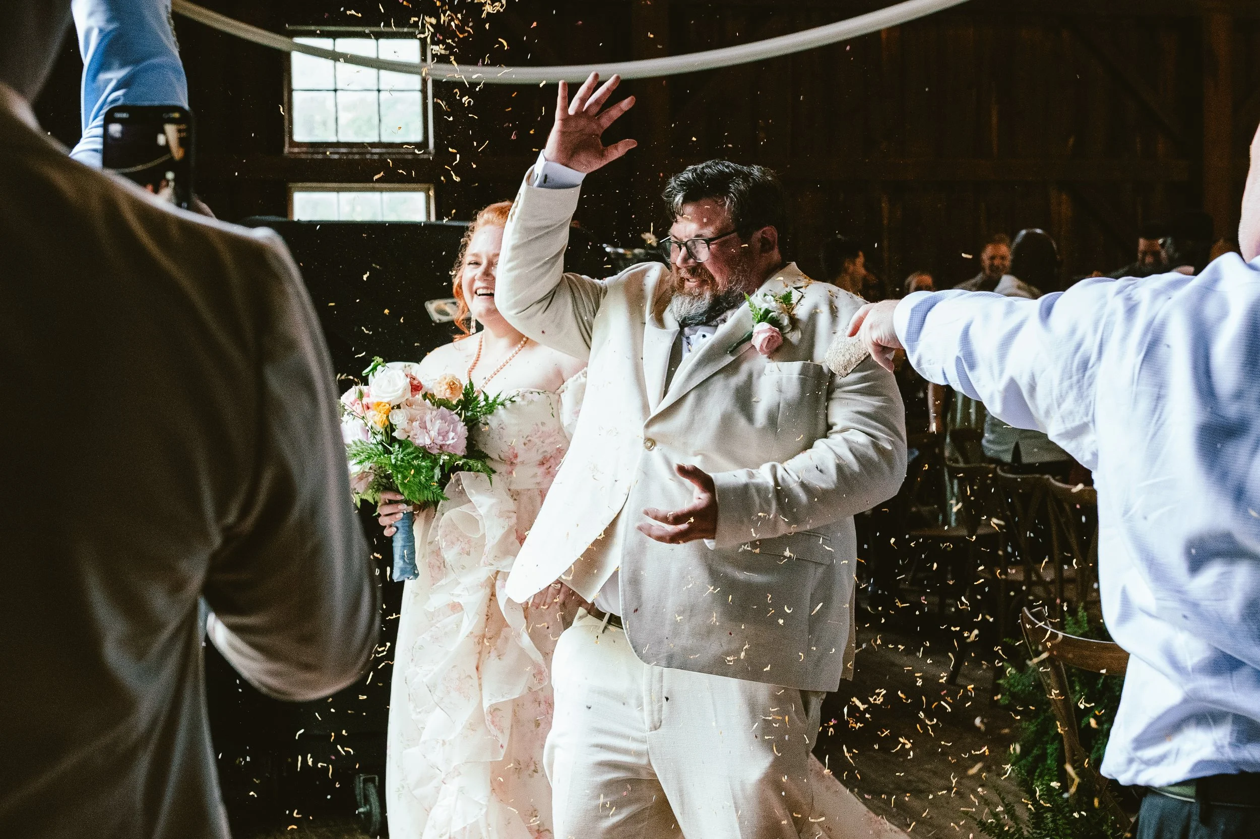 A joyful wedding celebration with a bride in a floral dress holding a bouquet and a groom in a cream suit throwing confetti in a rustic, wooden interior.