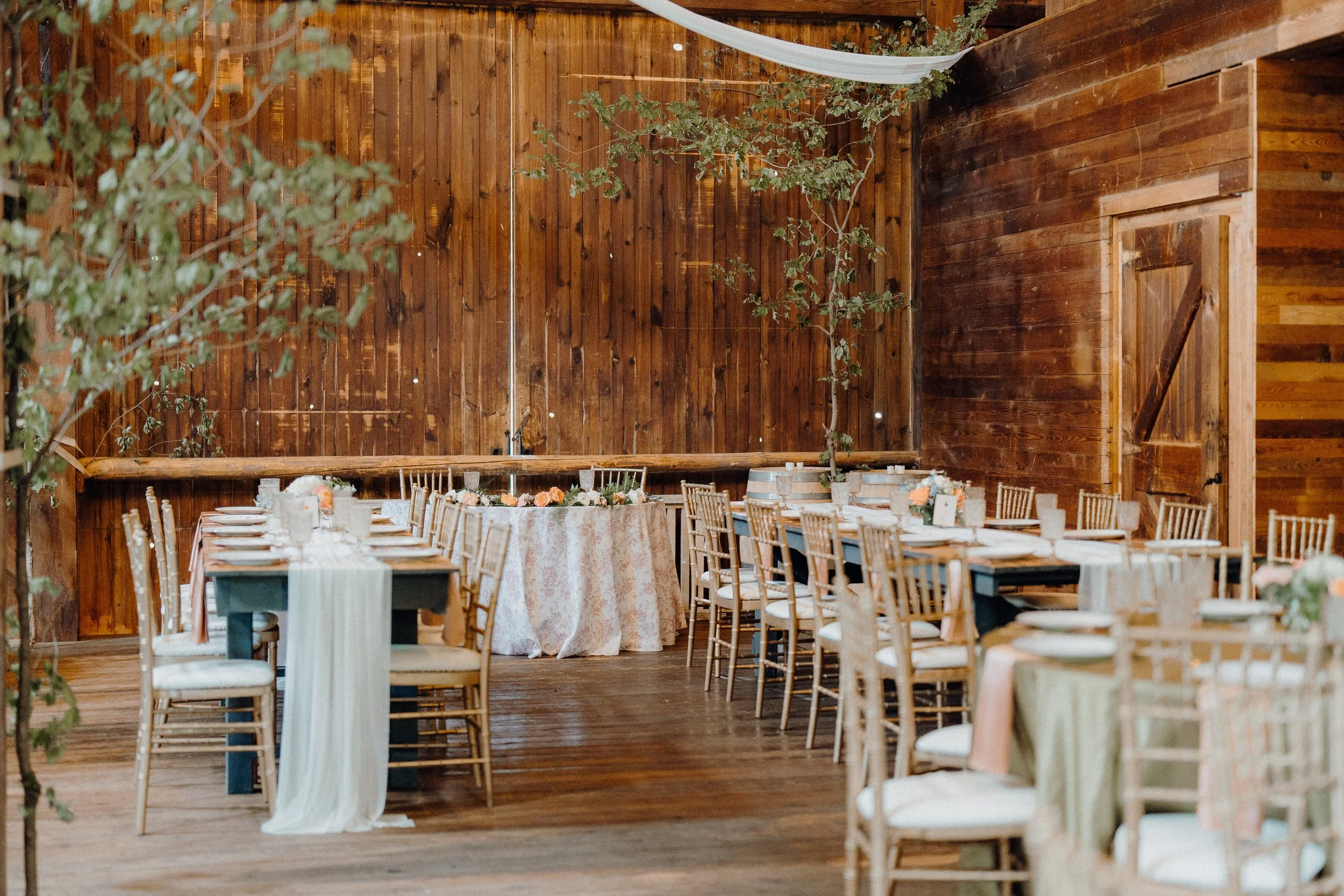 Decorated rustic event space with long wooden tables, gold chairs, floral centerpieces, and wooden walls, set for a celebration.