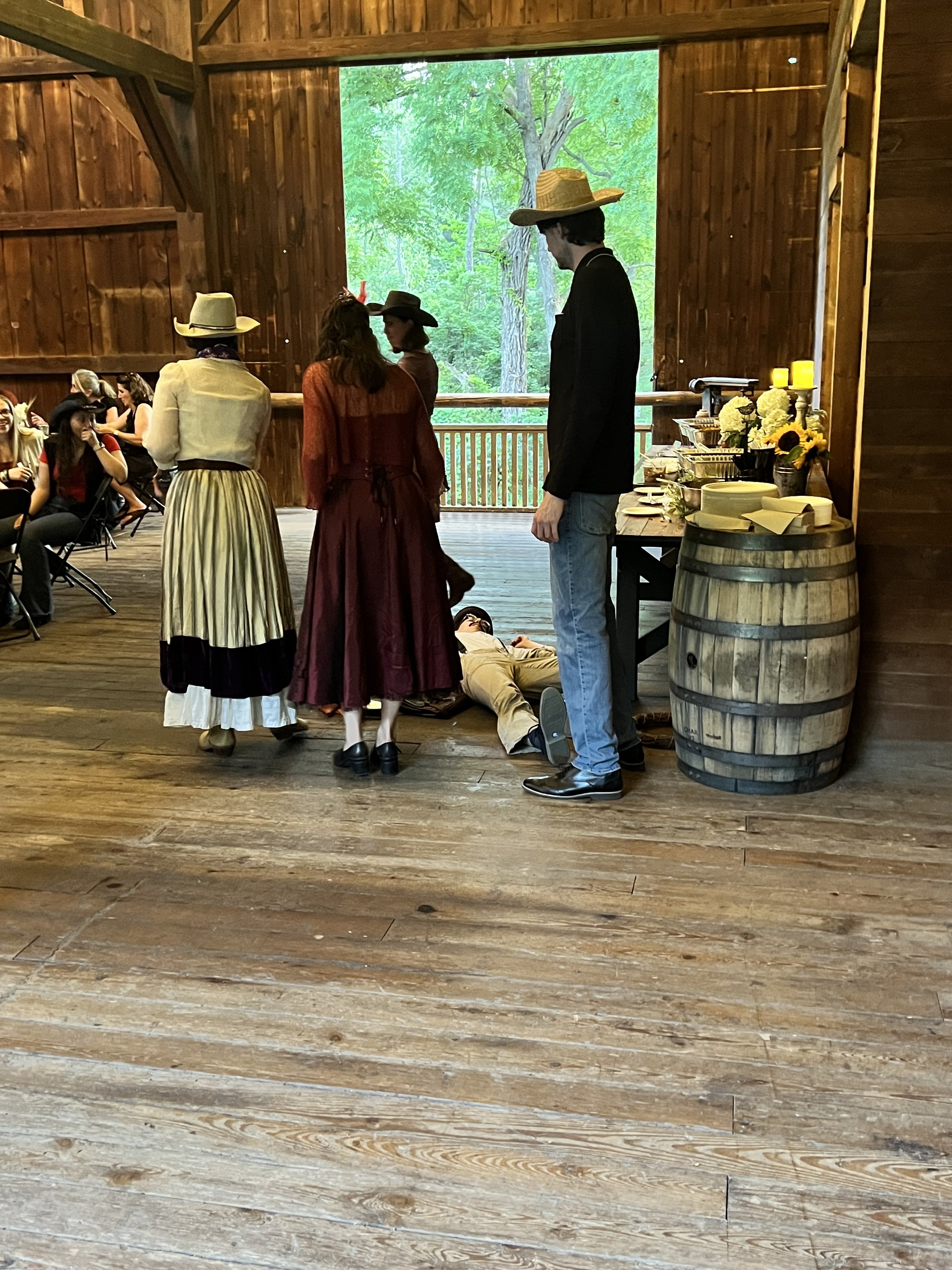 People in period costume gathered in a wooden barn setting with a person lying on the floor. They are wearing hats and western-style clothing.