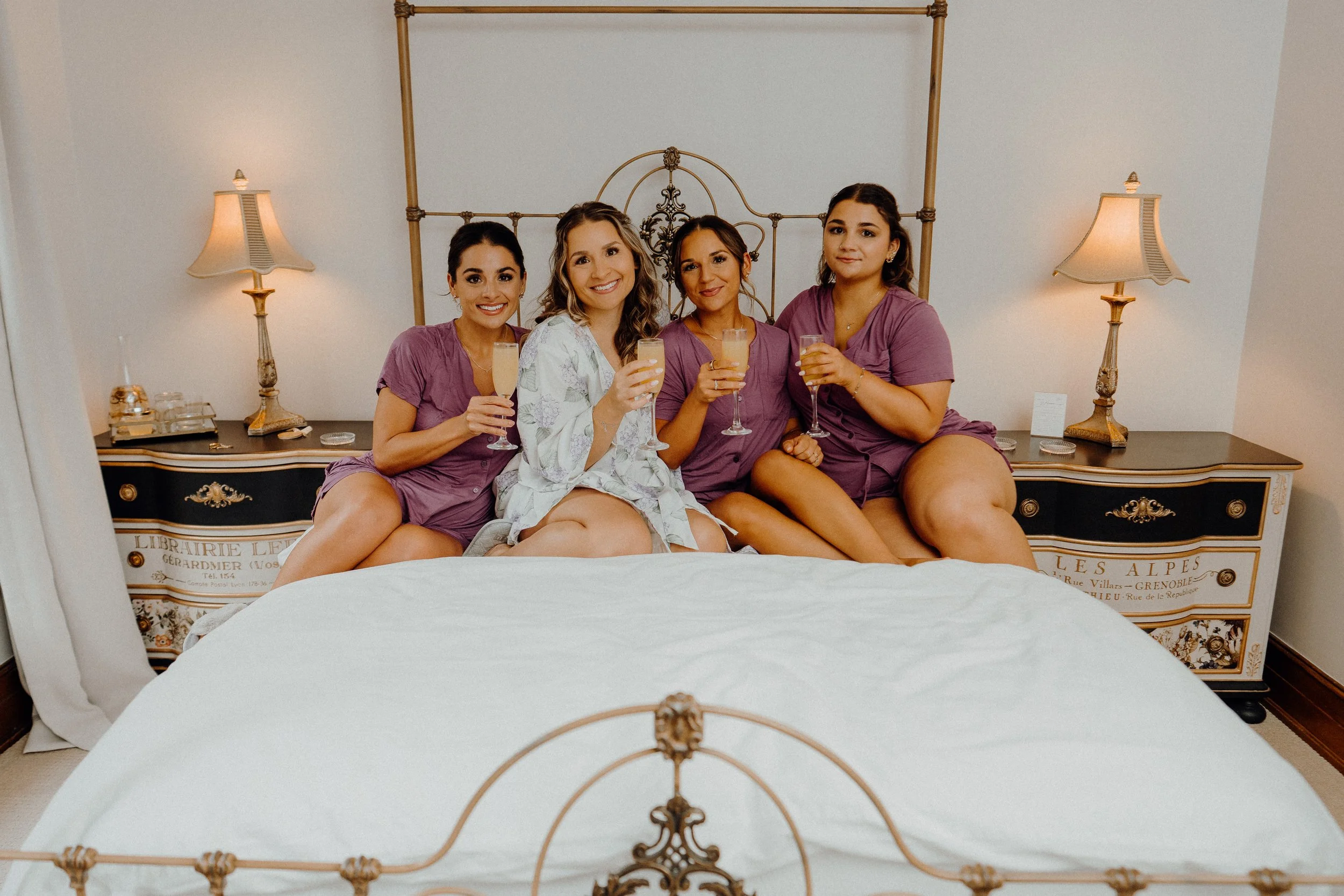 Four women sitting on a bed, smiling and holding glasses of champagne, in a bedroom with two nightstands and matching lamps.