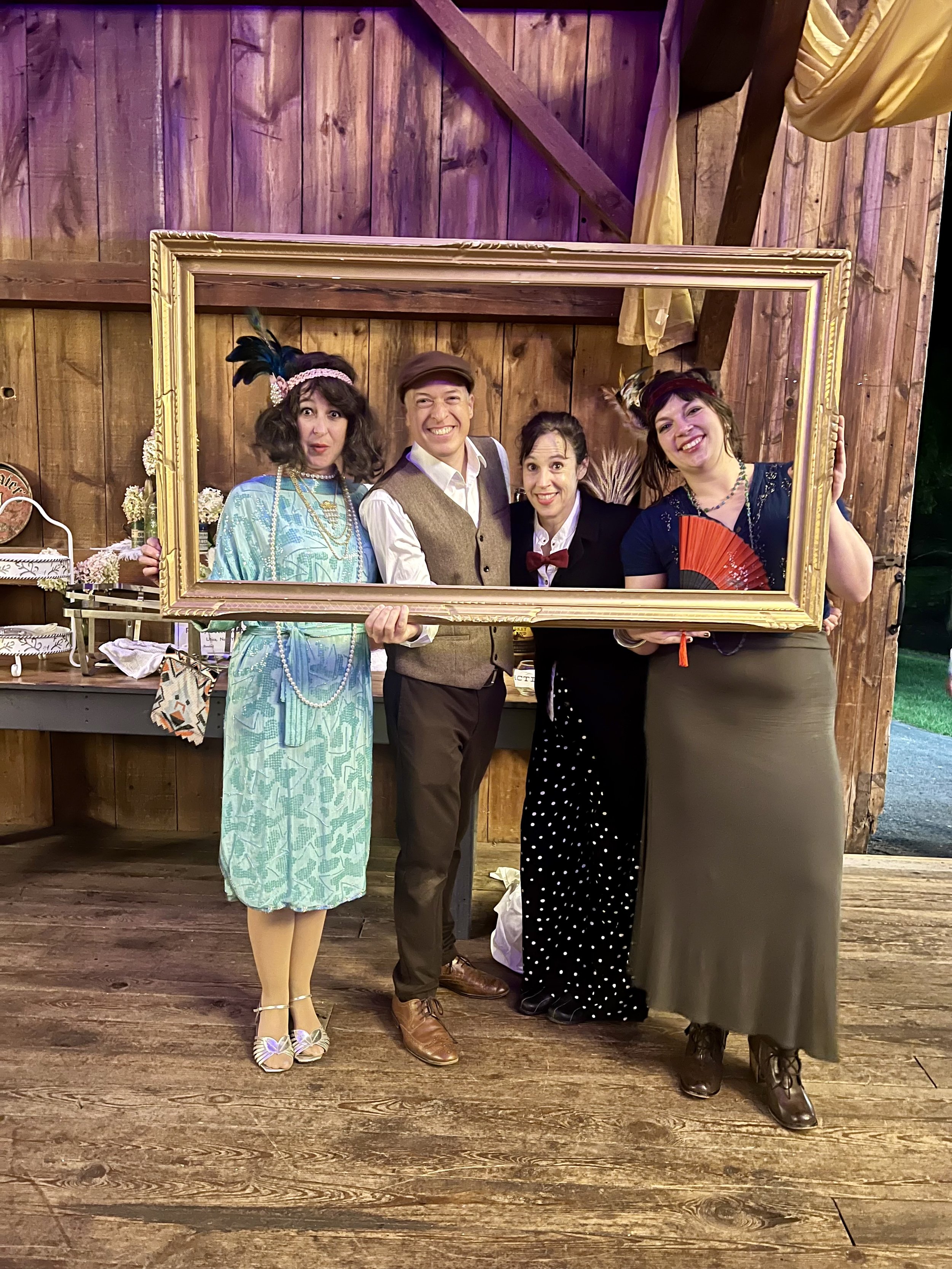Group of four adults in vintage attire posing with a large empty picture frame in a rustic wooden setting.