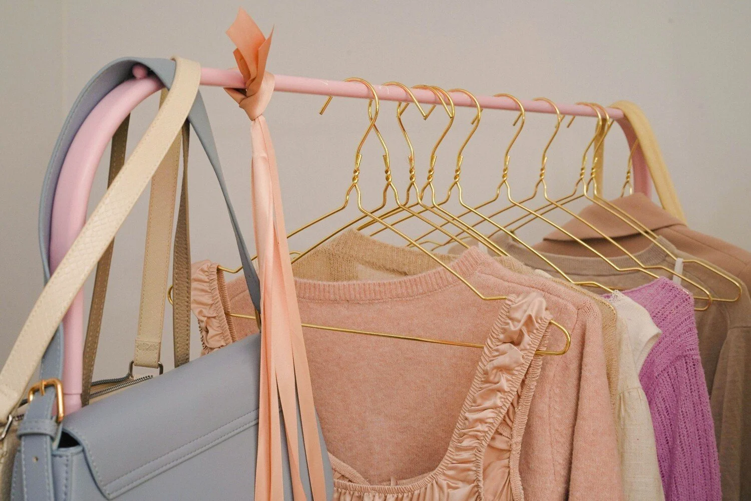 Clothes hanging on a pink clothing rack, including sweaters and tops in pastel colors, with some gold hangers and handbags hanging on the side.