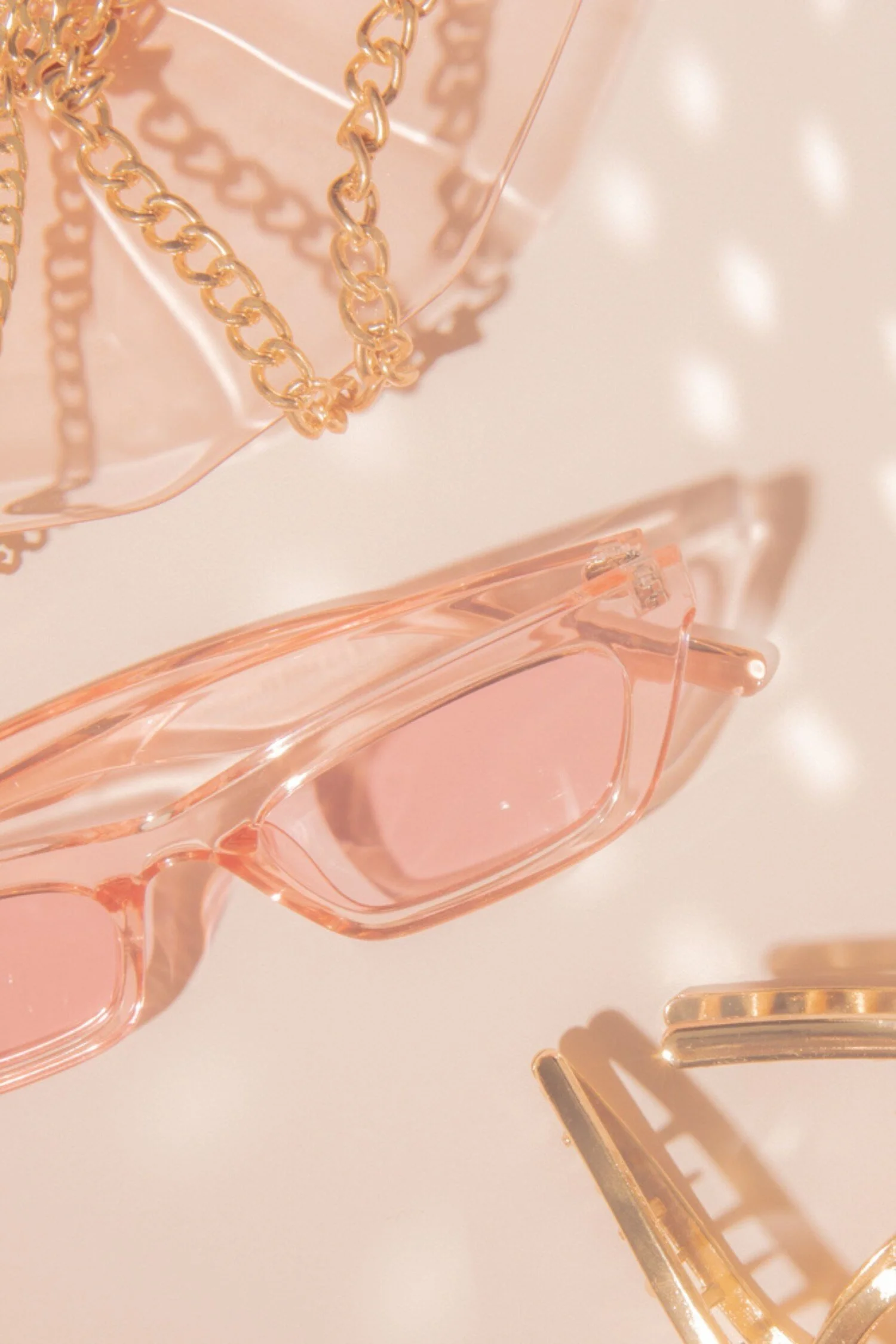 Pink tinted sunglasses, gold chain bracelets, and part of a gold clip or hair accessory on a light pink surface.