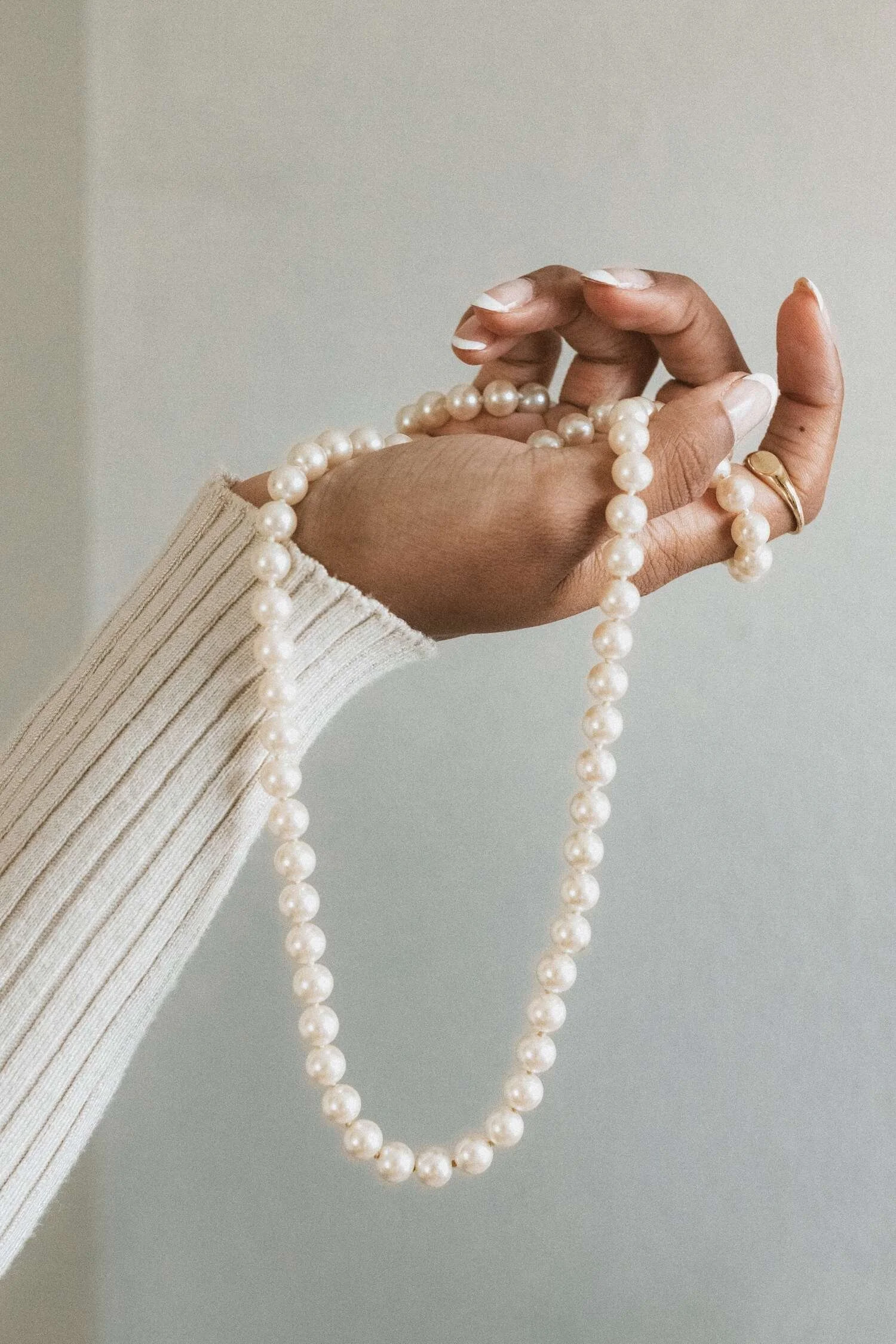 Close-up of a hand holding a string of white pearl necklace, with the person wearing a cream or beige ribbed sweater and gold rings.