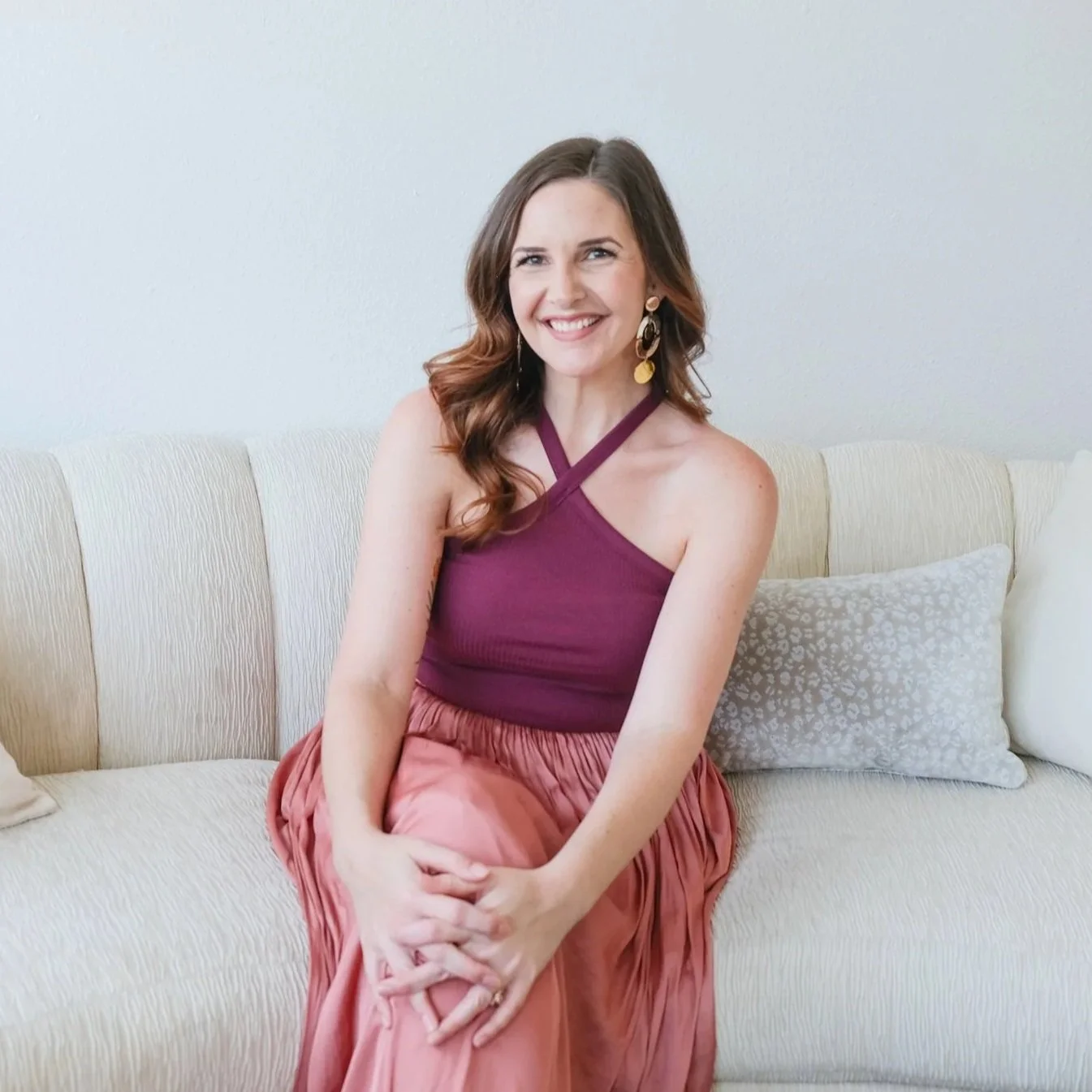 A woman with long wavy brown hair, wearing a burgundy halter top and a pink pleated skirt, sits on a beige couch and smiles at the camera.