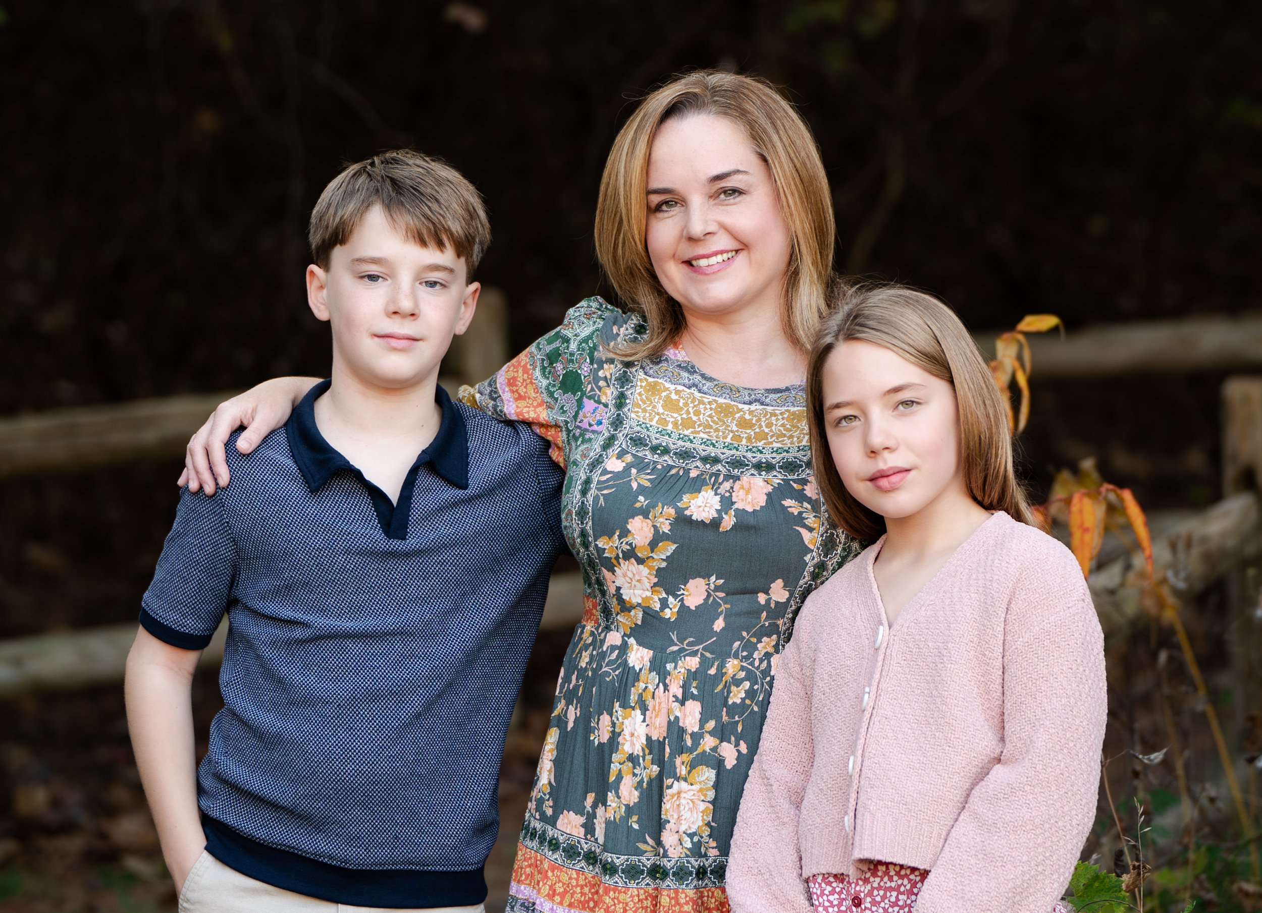 Woman and two children posing outdoors in front of a wooden fence, with trees in the background.
