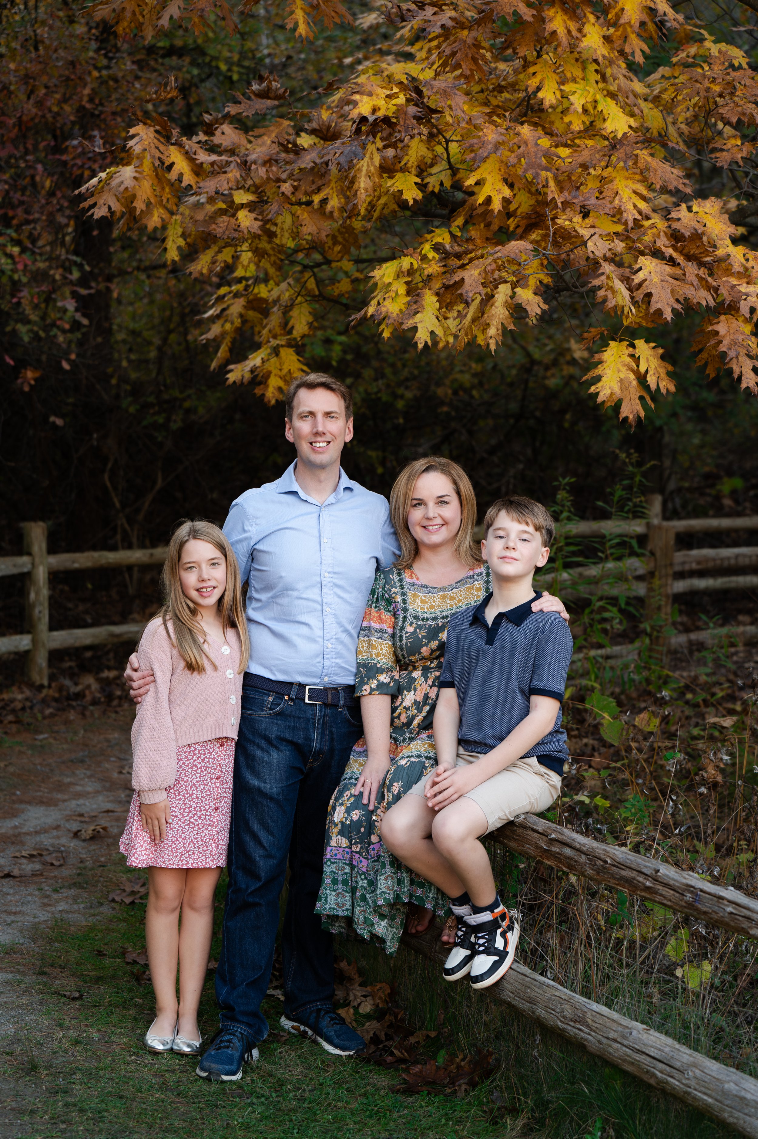 Family of four posing outdoors in autumn, with colorful fall leaves and wooden fence in background.