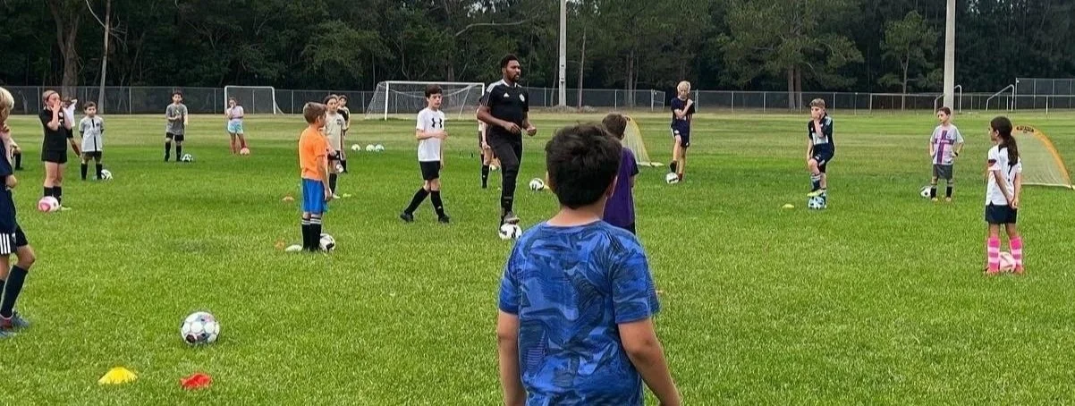 Children and a coach on a soccer field participating in a training session. The children are standing with soccer balls, with some lining up or listening to instructions, on a grassy field with goal nets in the background.