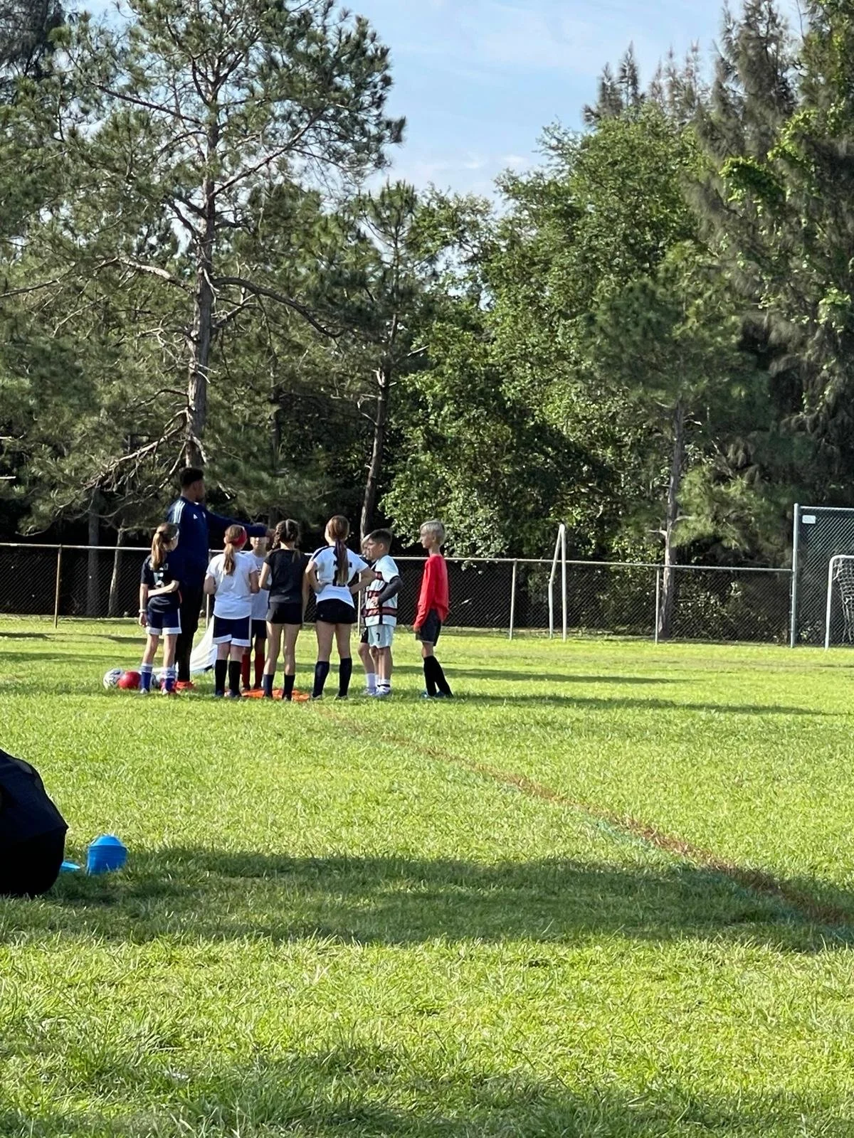 Group of young children and a coach on a soccer field during practice, with trees and a fence in the background.