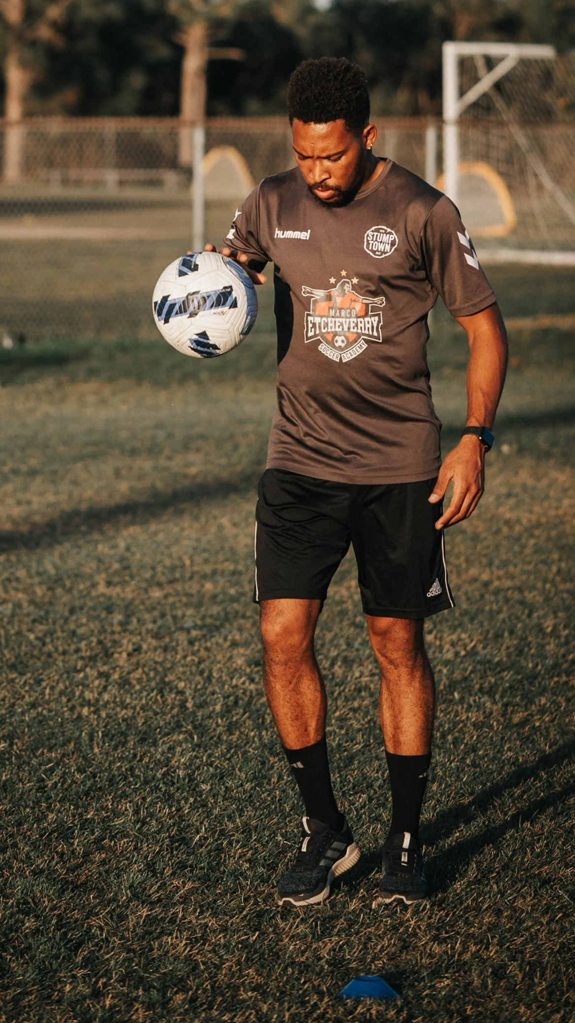 A man standing on a grassy soccer field, holding a soccer ball, wearing a dark gray sports jersey, black shorts, black socks, and black athletic shoes, with goalposts and a chain-link fence in the background.