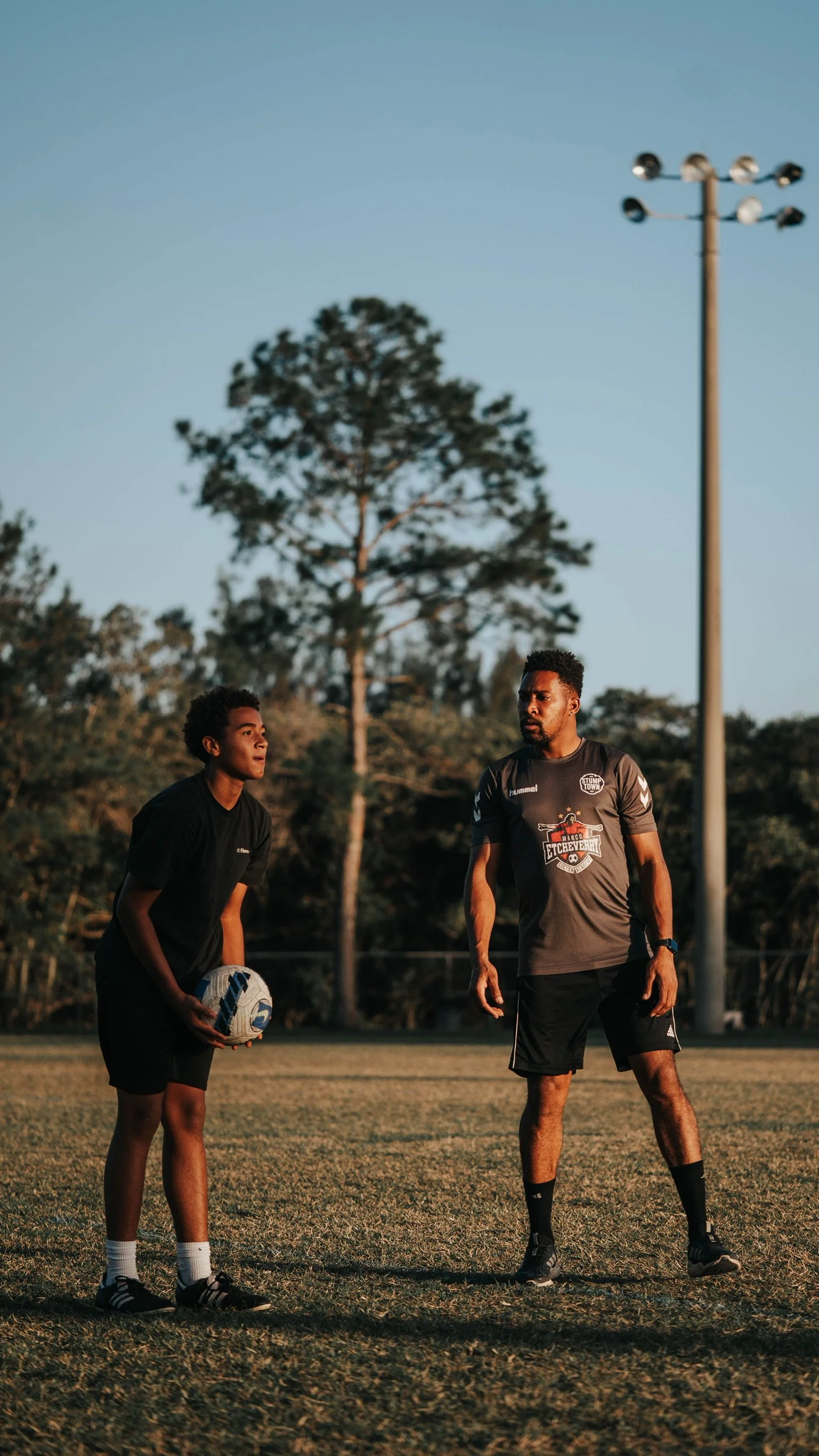 A young boy and a man standing on a grassy field during sunset, with trees and a tall light pole in the background, preparing for a soccer practice or game.