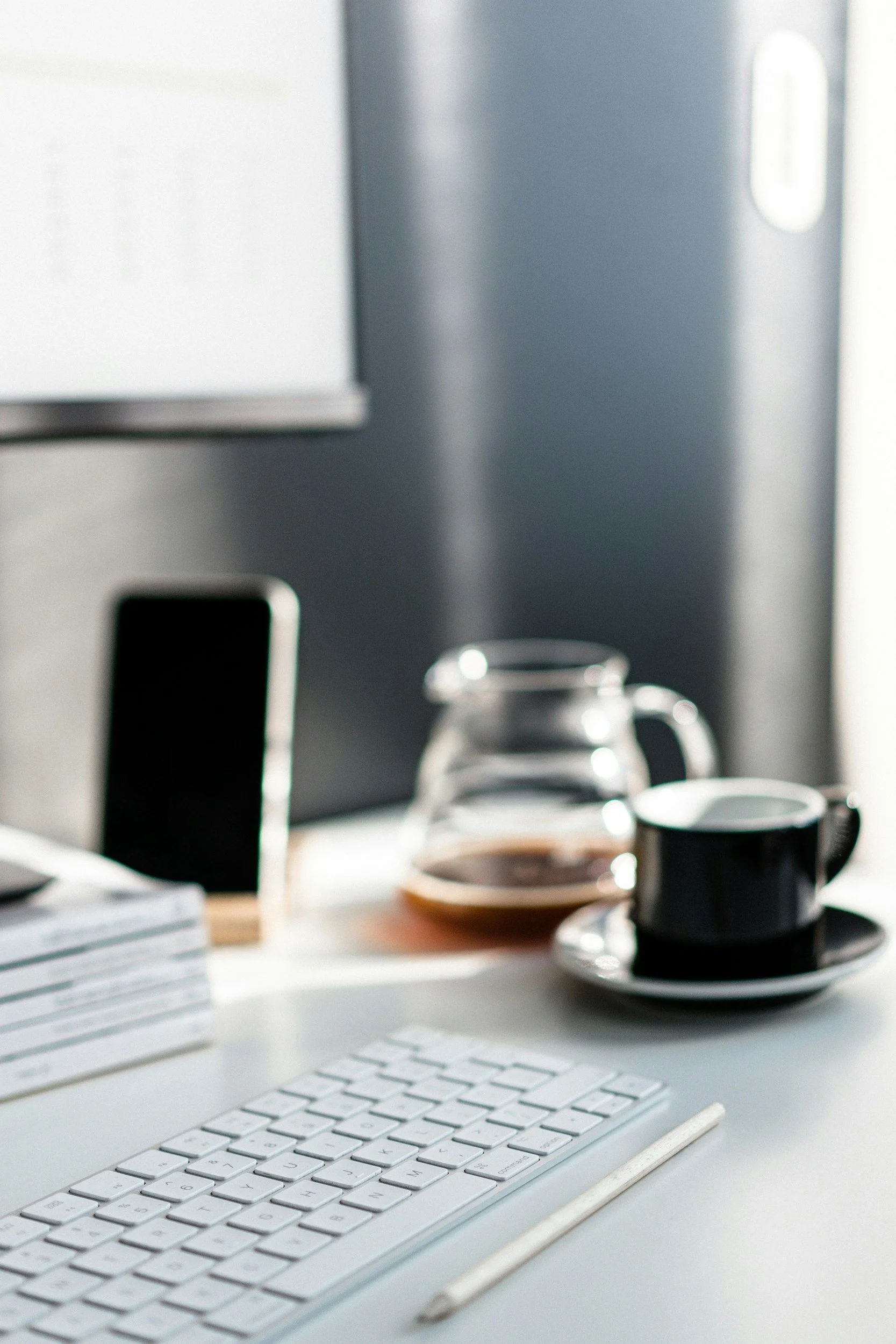 Computer desk with white keyboard, desk calendar, and pencil in foreground. In the background, a glass coffee pot, a black coffee cup on a saucer, and a smartphone standing upright on the desk, with a computer monitor partially visible.
