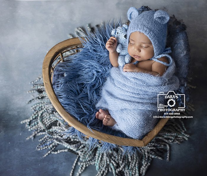 A newborn baby peacefully sleeping in a heart-shaped wicker basket, wrapped in light blue blankets, wearing a blue knit hat with bear ears, and holding a small stuffed bear. The basket is lined with a fluffy blue blanket and rests on a patterned blac