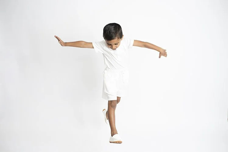 A young girl in a white outfit balancing on one foot with arms extended against a plain white background.