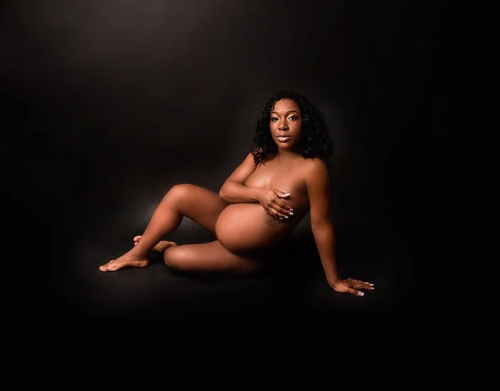 A pregnant woman with dark curly hair, sitting on a black background, covering her breast with her left hand and resting her right hand on the ground.