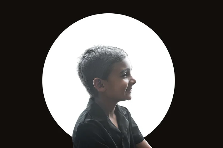 Profile of a young boy with short hair smiling, in front of a circular white background with a black surrounding.