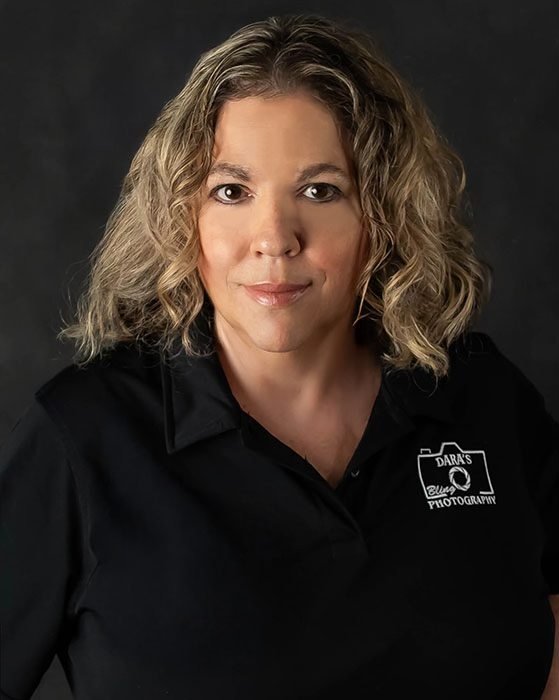 Portrait of a woman with shoulder-length wavy blonde hair, wearing a black polo shirt with a logo that says "Dara's Photography." She is smiling slightly against a dark background.