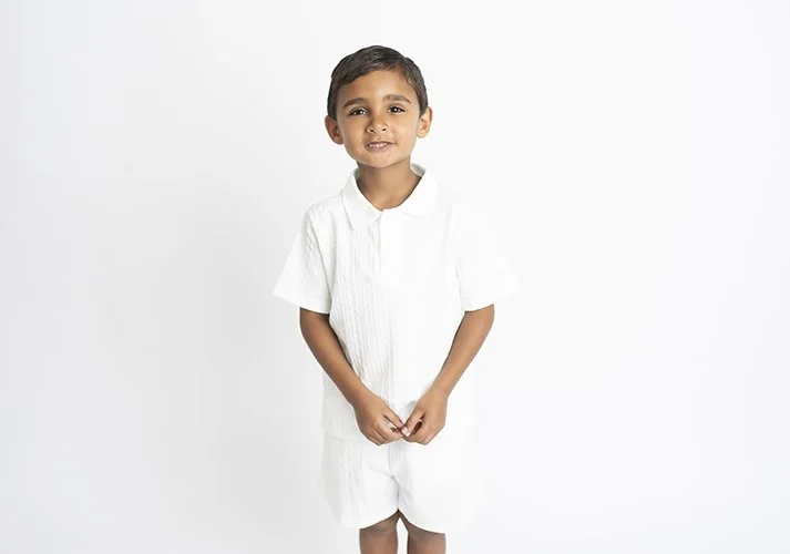 Young boy smiling, standing against a plain light background, wearing a white shirt and white shorts.