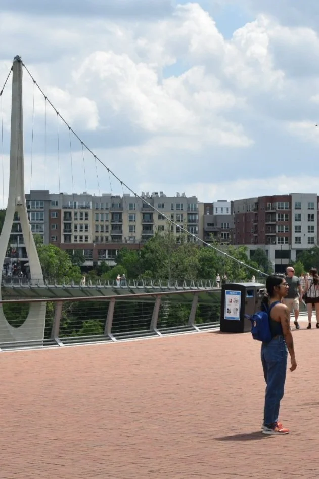 People walking on a scenic bridge with modern buildings in the background, capturing a lively urban atmosphere on a sunny day.