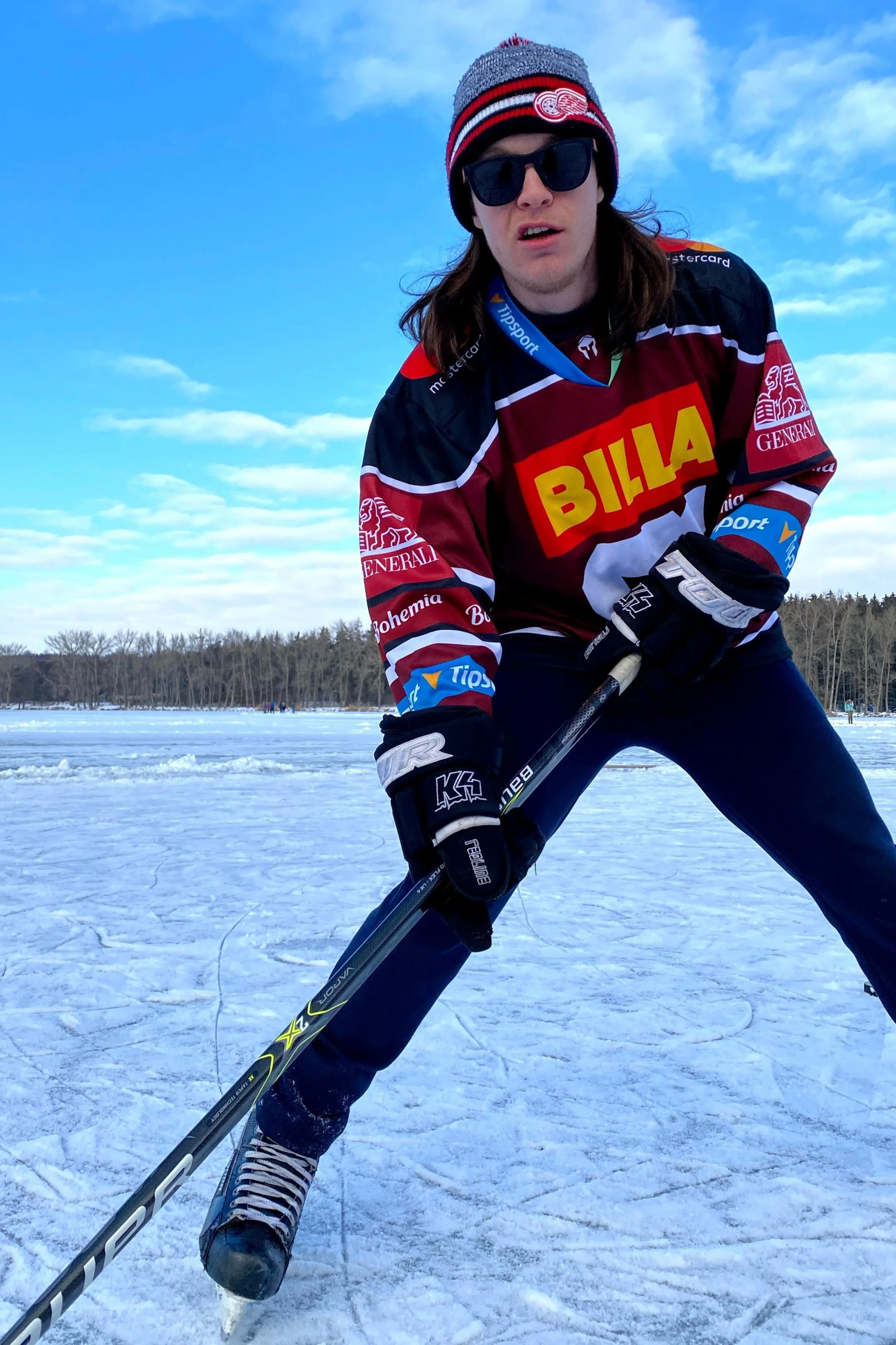 Bob Ruediger wearing ice skates, a hockey jersey and gloves, sunglasses, and a beanie, poses on a frozen lake with a hockey stick, ready to play. The background features a clear blue sky and a forest.