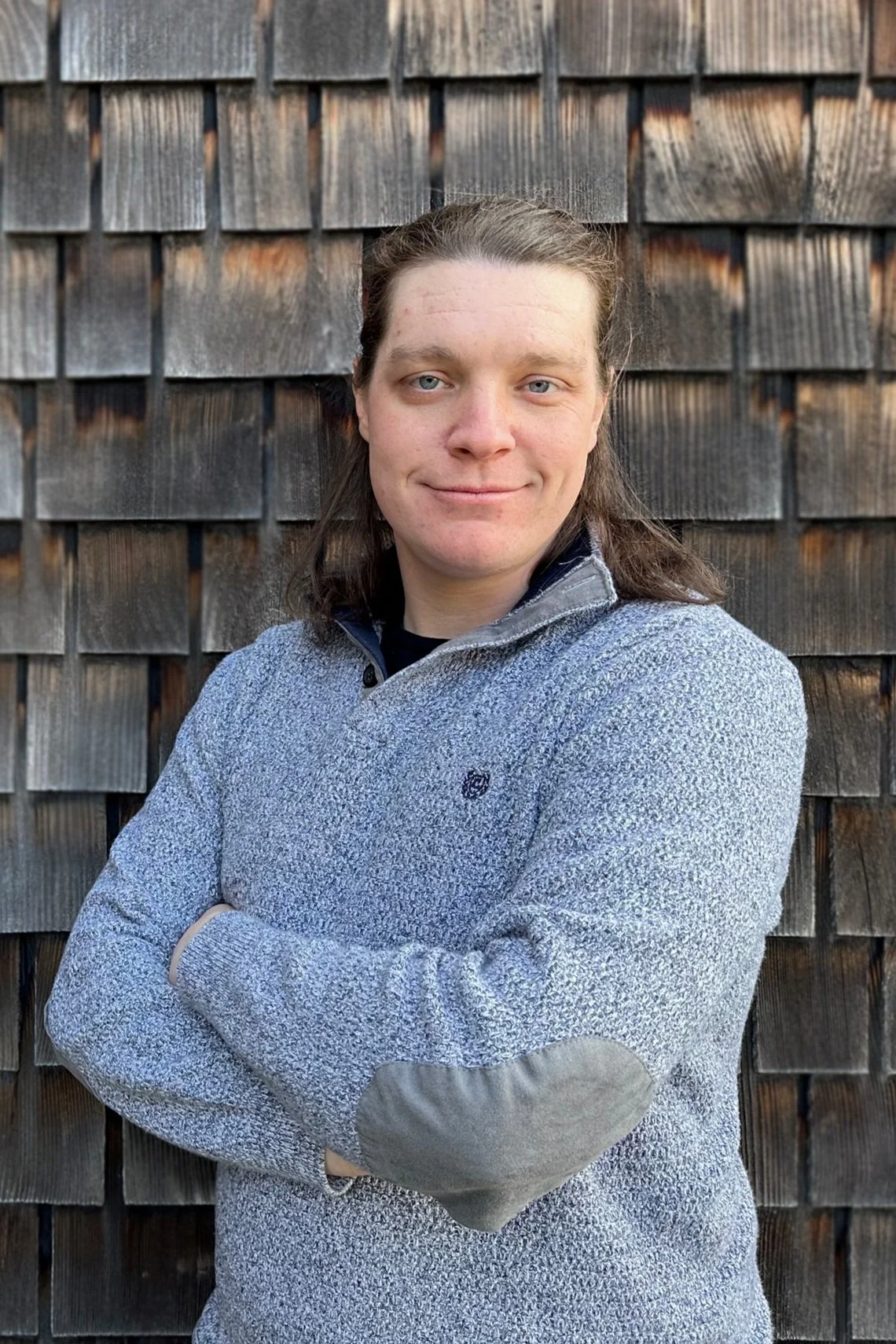 Bob Ruediger stands smiling with his arms folded in front of a wooden background.