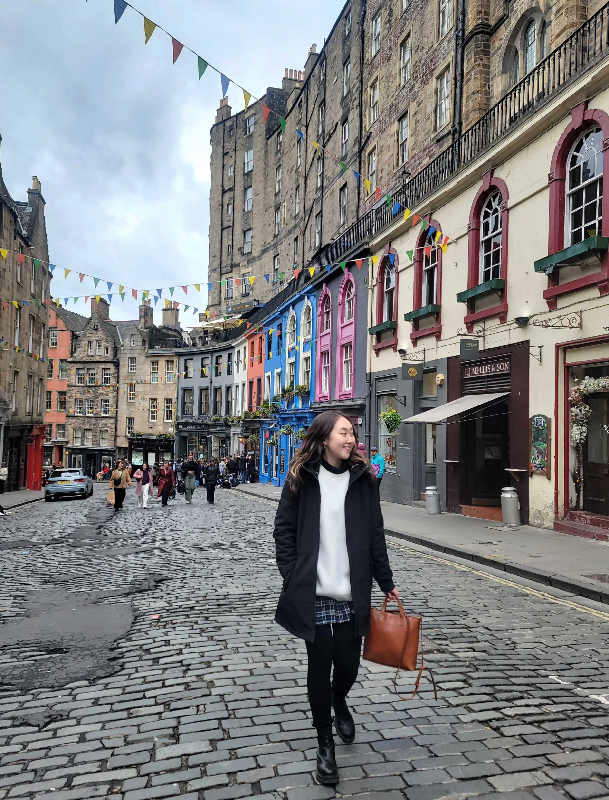 Julie Kim walking down a colorful, cobblestone street in Edinburgh, with colorful buildings and festive flags overhead.