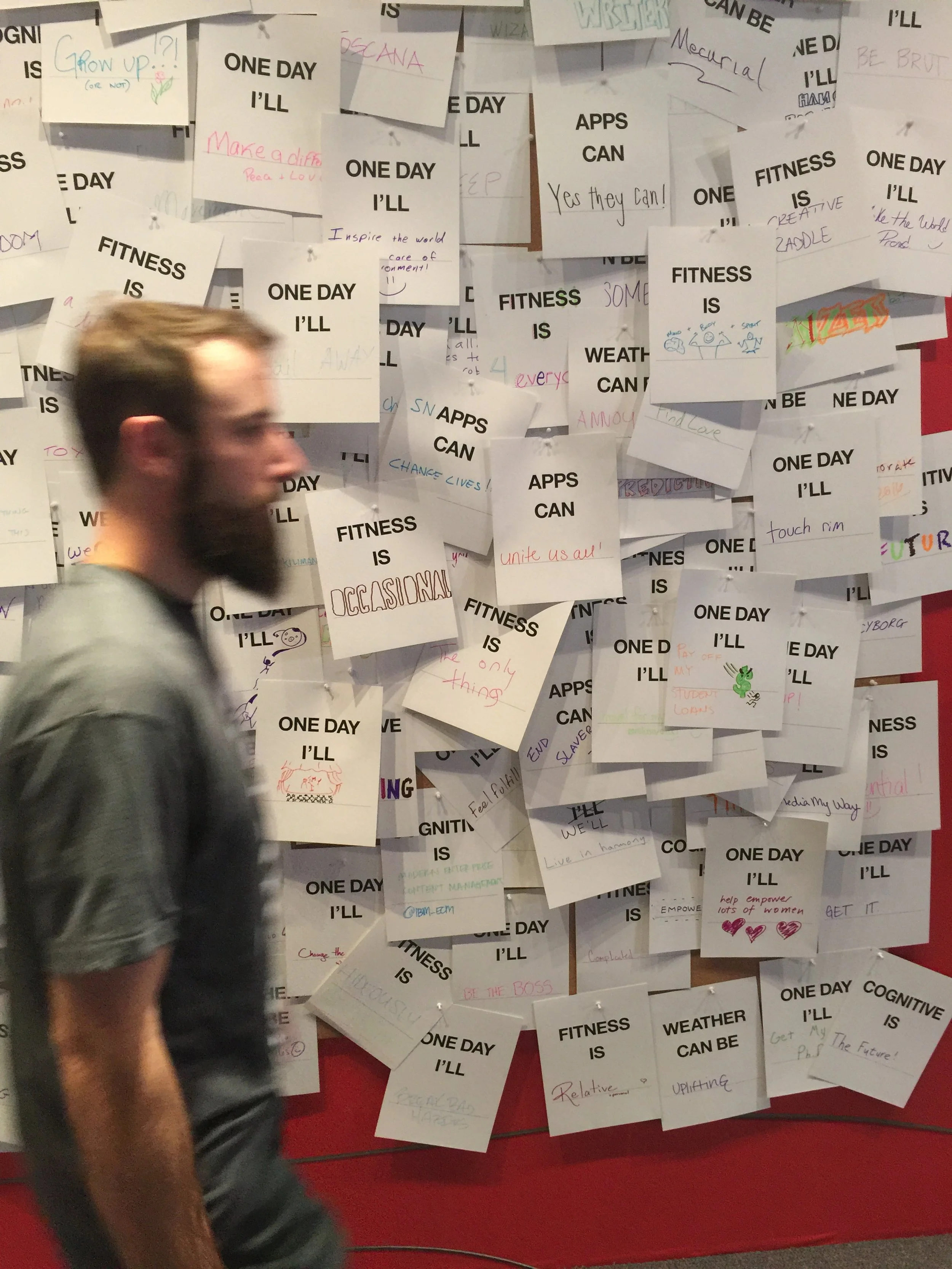 A man walks past a wall covered in various notes and messages. Each note has a printed statement starting with "One day I'll," "Apps can," "Fitness is," and "Weather can be," followed by handwritten responses in different colors.