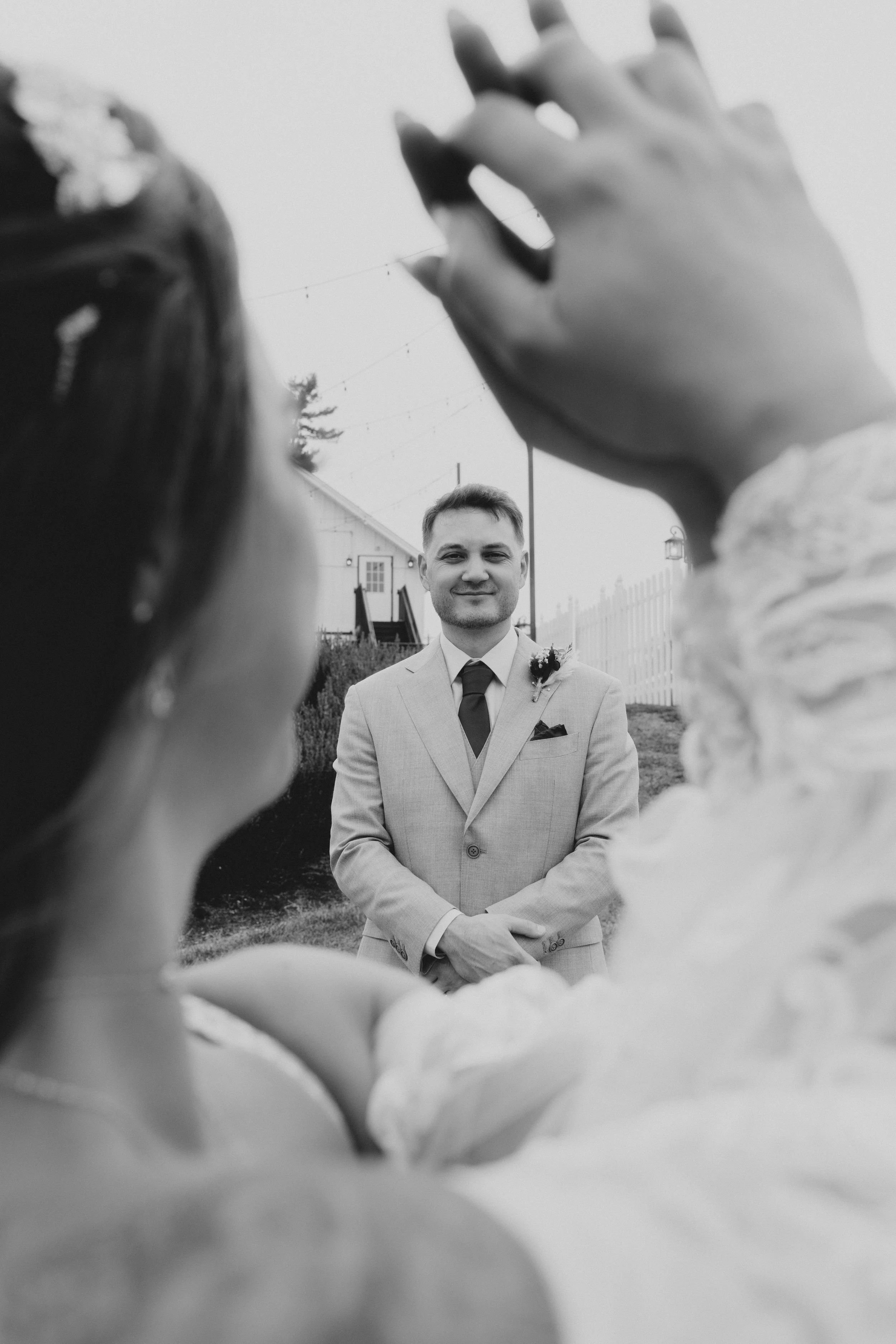 A black and white photograph of a couple, with the man standing in the background and the woman in the foreground, capturing a moment outdoors. The man is dressed in a suit, smiling, with his hands clasped. The woman's face is partially visible and s