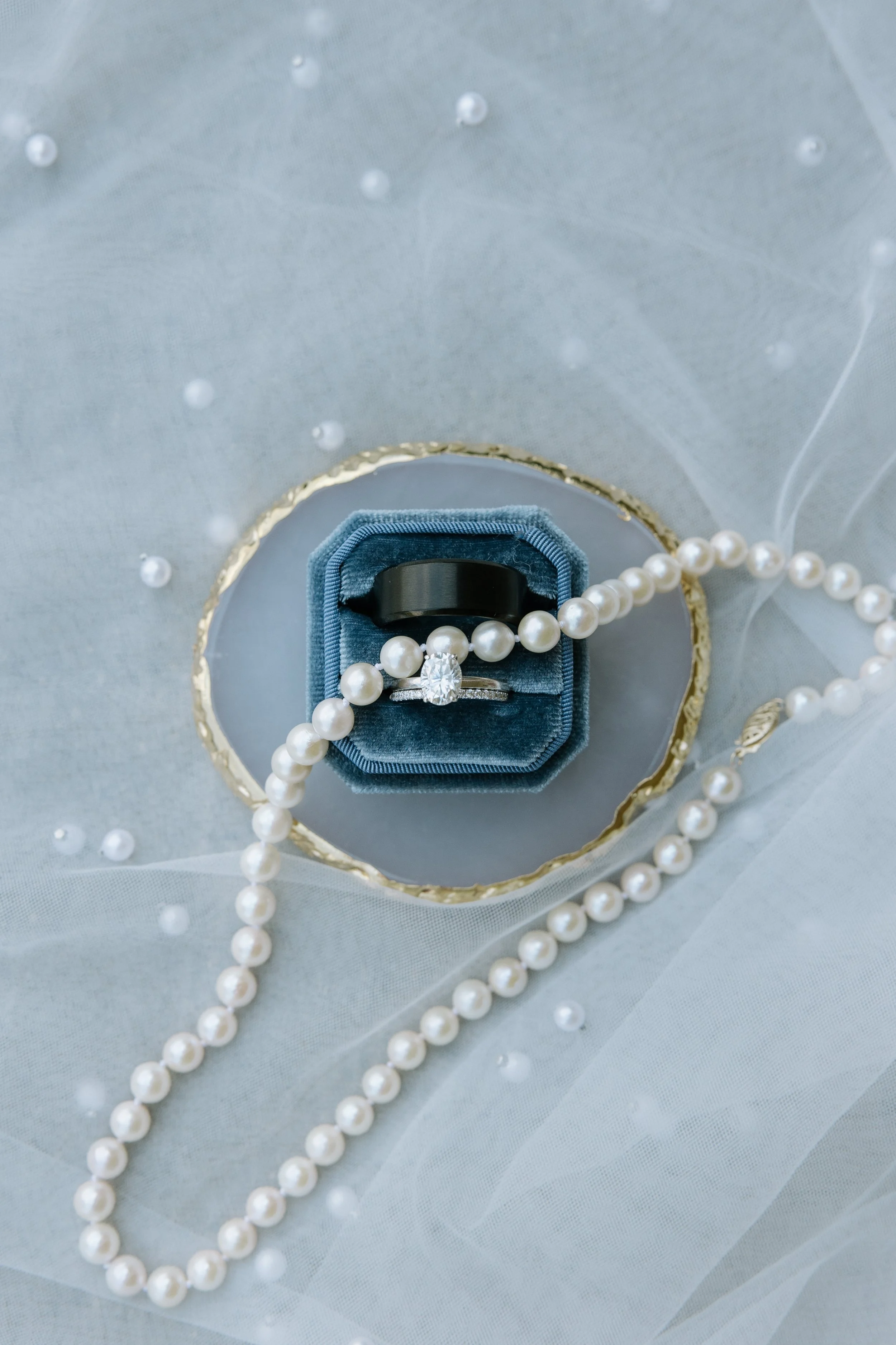 Close-up of a jewelry display with an engagement ring and wedding band, surrounded by a pearl necklace and pearls on a decorative fabric background.
