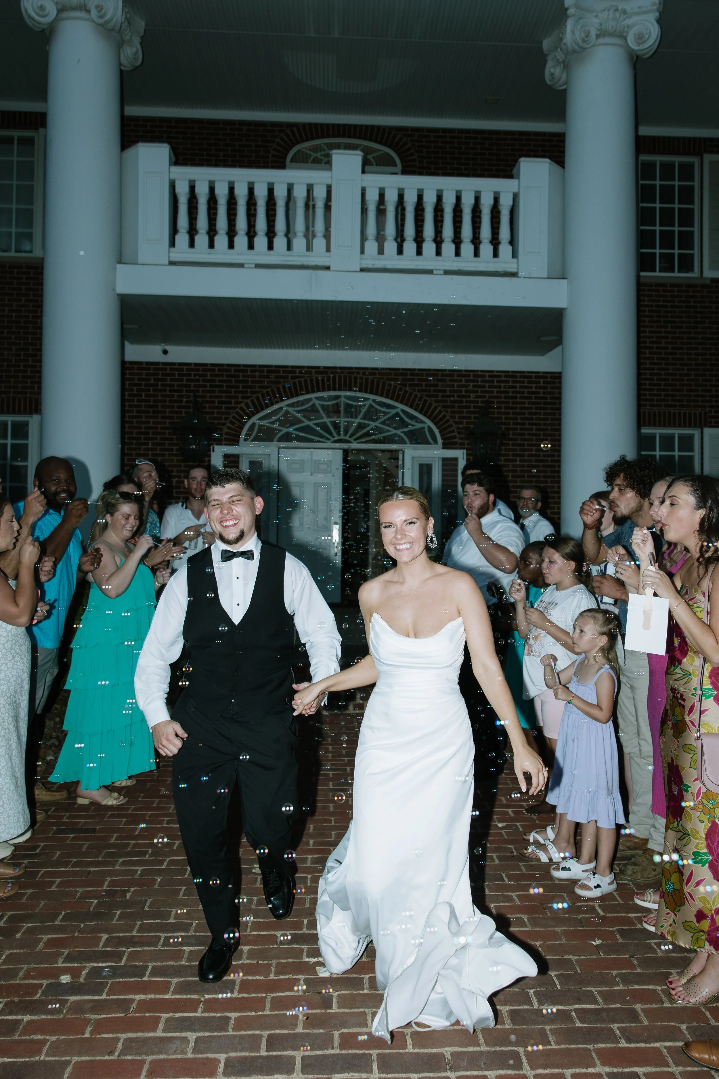 A bride and groom smiling and walking hand-in-hand through a crowd of friends and family, celebrating their wedding outside a venue with white columns and a brick facade at night.