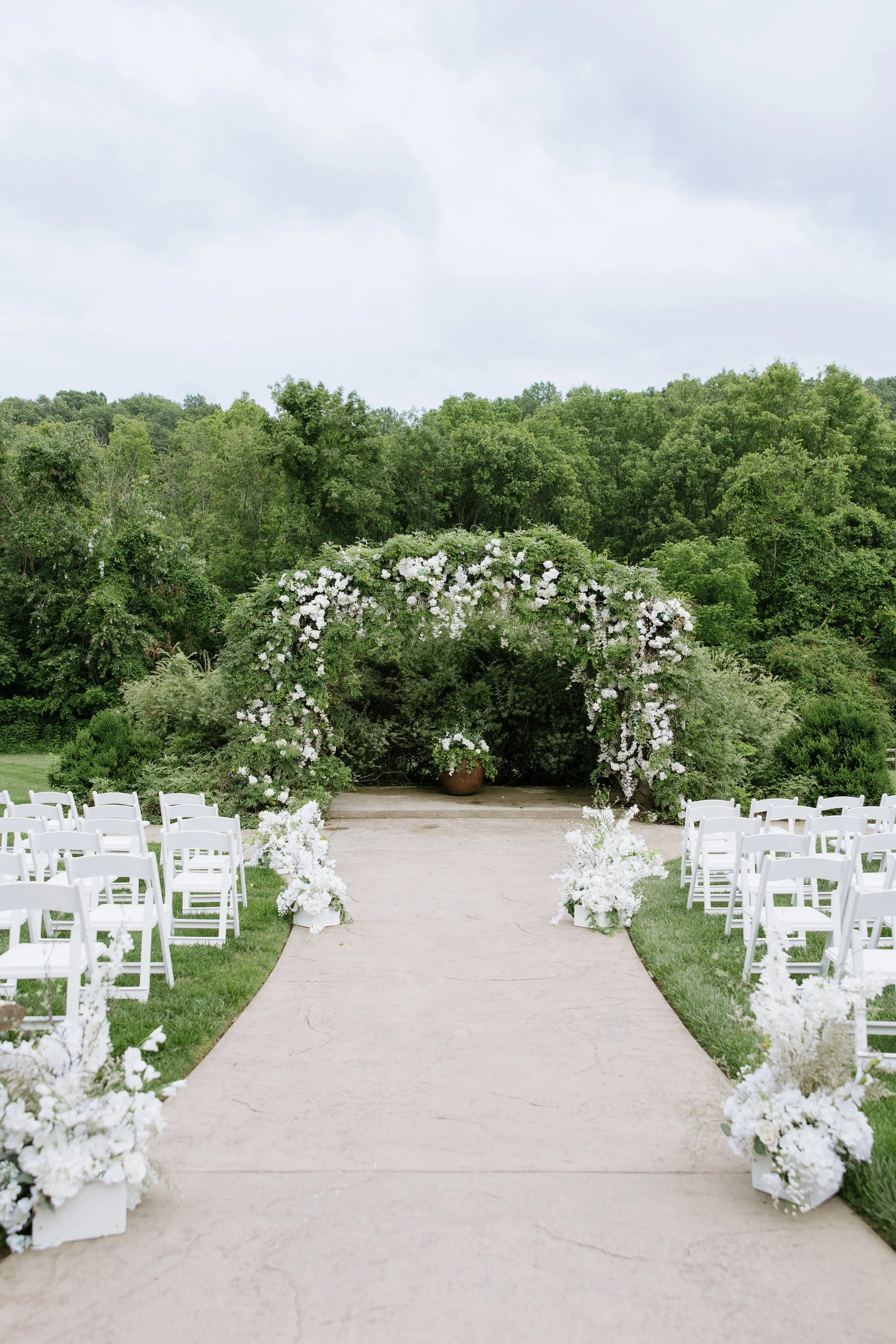 Outdoor wedding ceremony setup with white chairs arranged on a grassy area, white floral arrangements on the ground, and a floral arch at the end of the aisle, against a background of green trees under a cloudy sky.