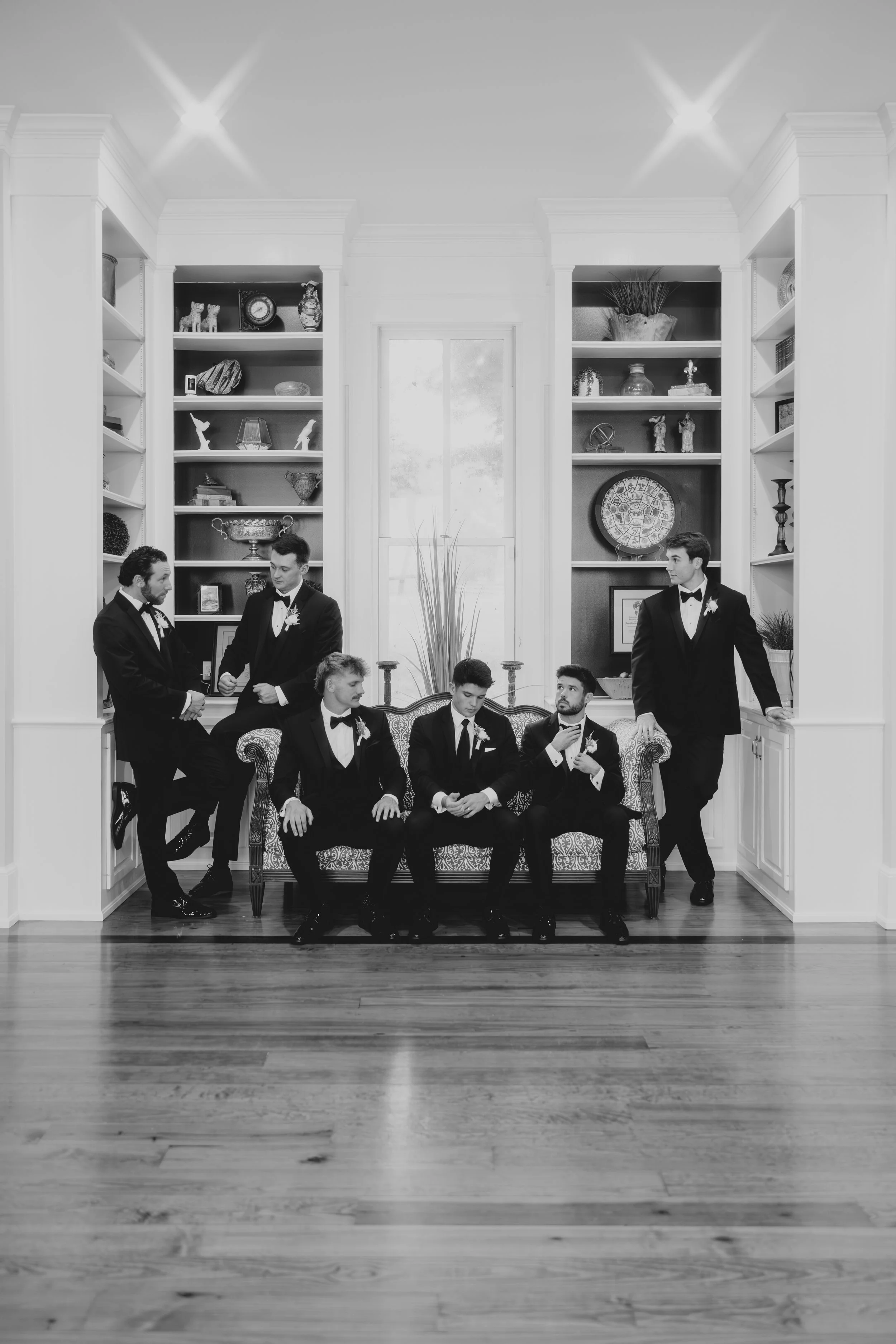 A group of six men in tuxedos sitting and standing on and around a vintage sofa in a bright room with built-in bookshelves, window, and hardwood floor.