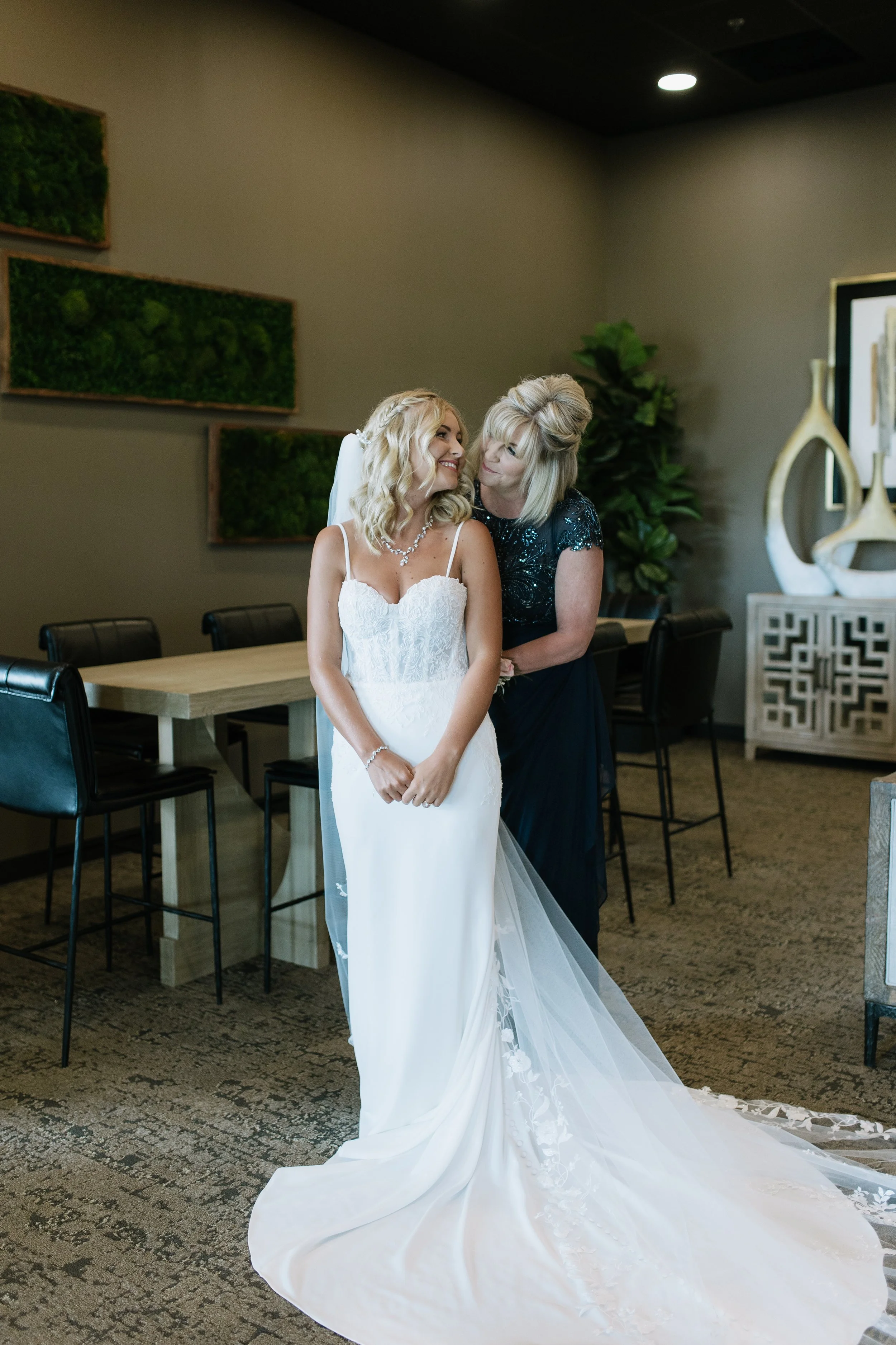A bride in a wedding dress smiling and looking at a woman, possibly her mother, standing behind her.