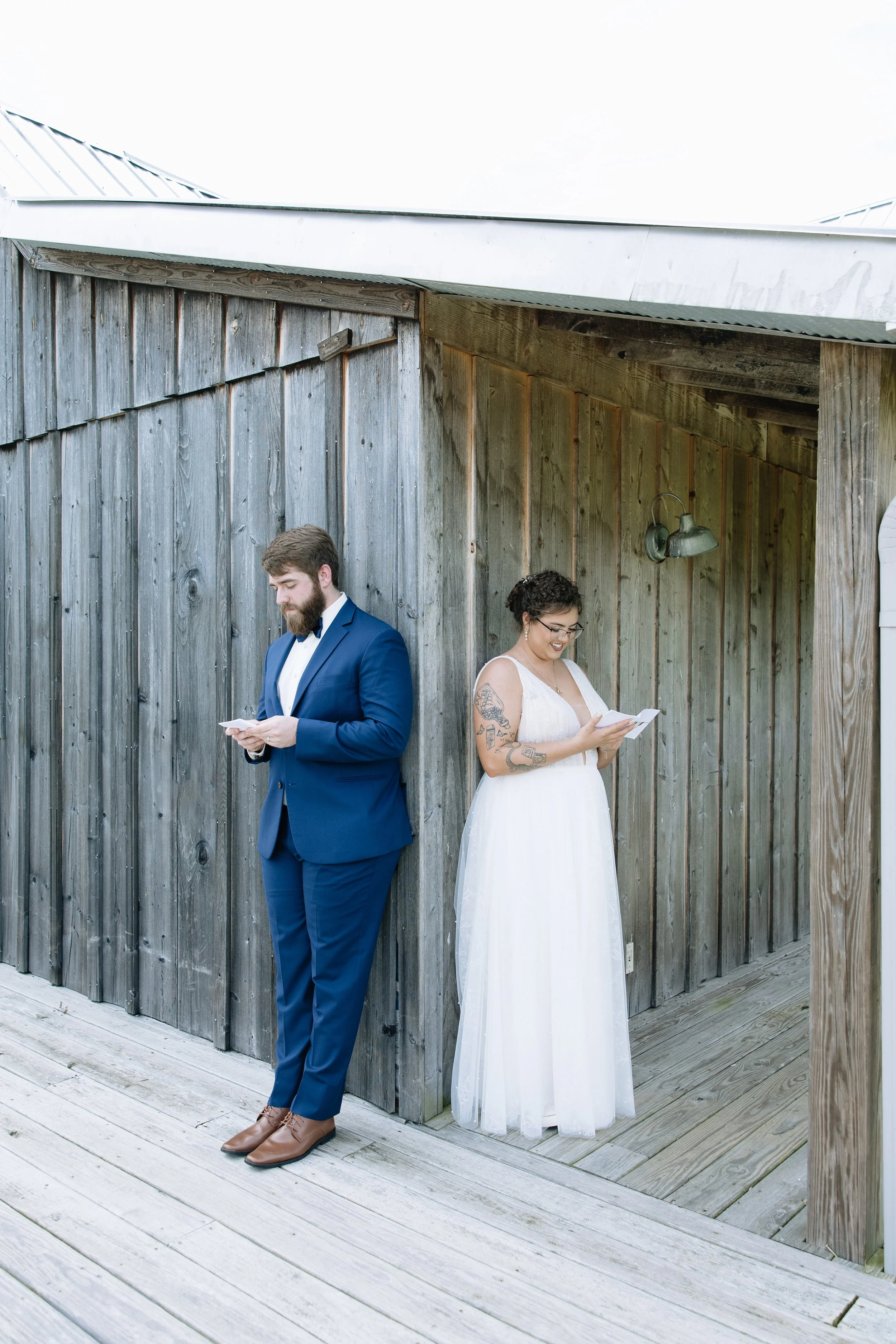 A man in a blue suit and a woman in a white dress standing against a wooden wall, both reading papers, outside on a wooden deck.
