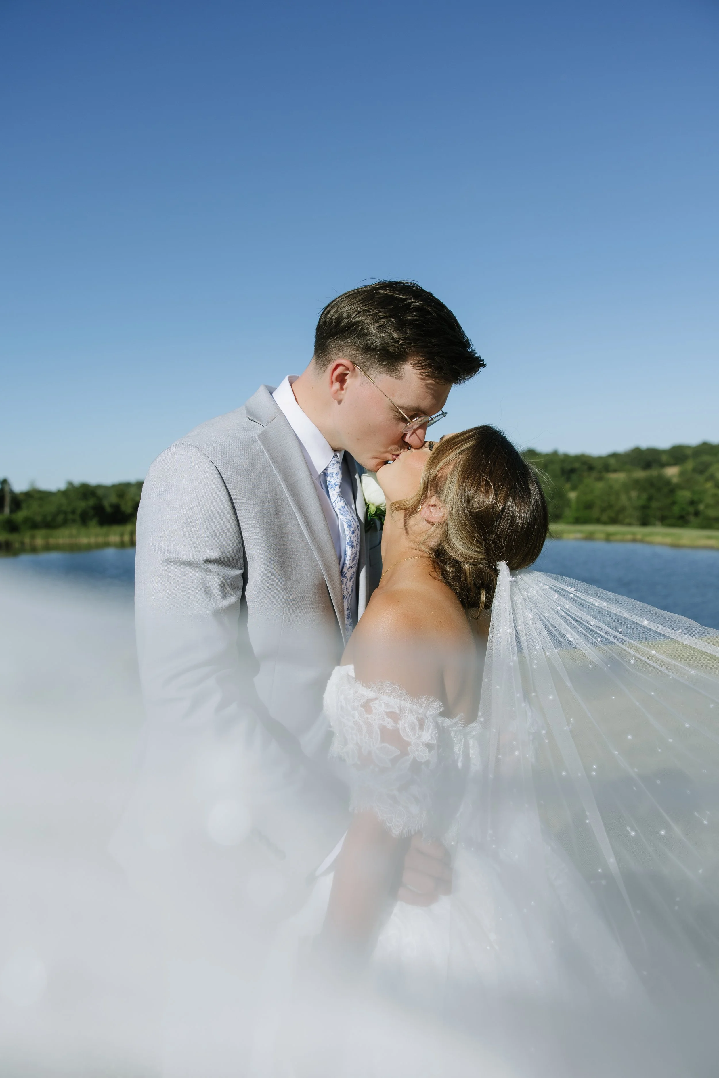 A man and woman in wedding attire sharing a kiss in an outdoor setting with trees in the background.