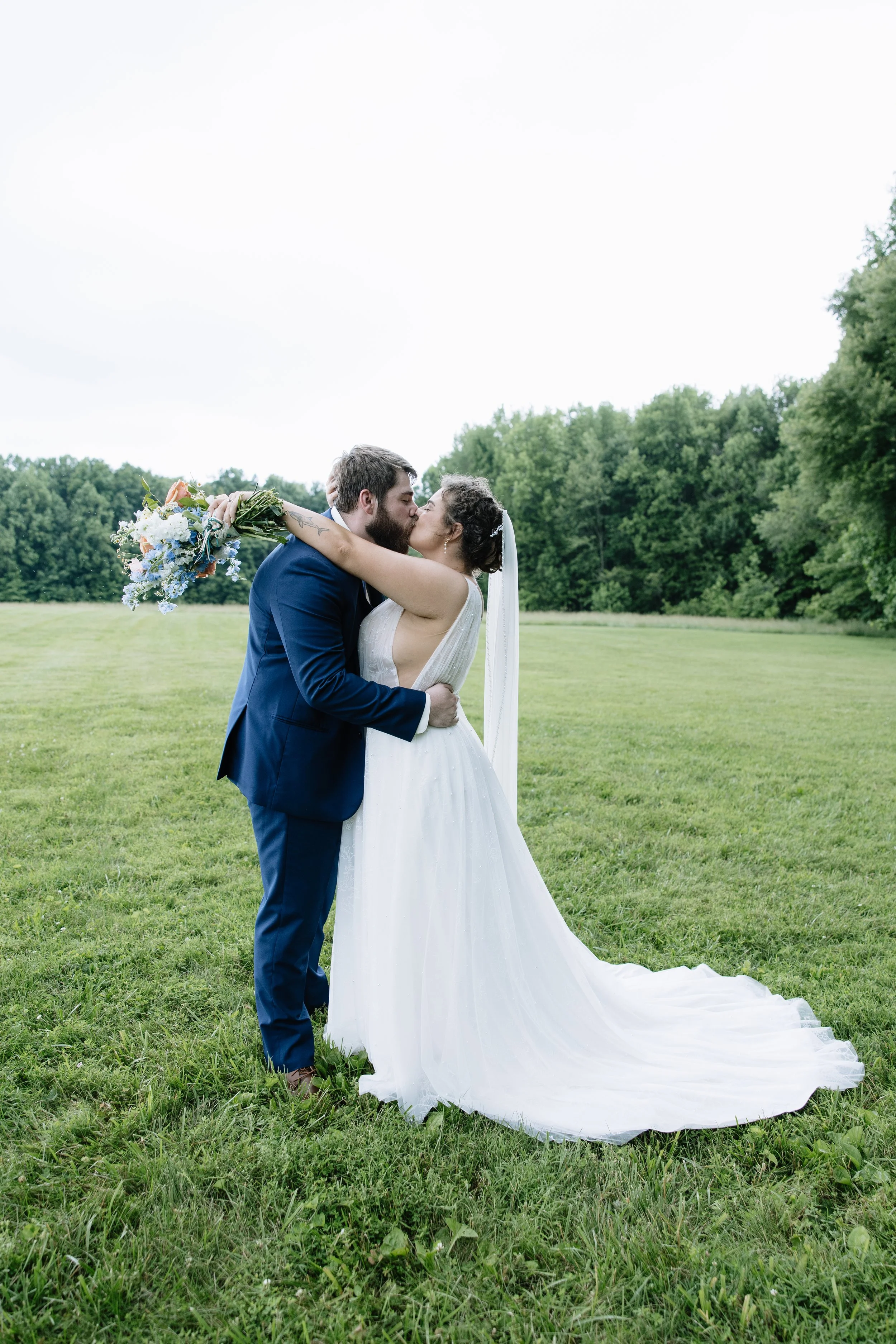 Bride in a white wedding dress and veil kissing groom in a blue suit holding a bouquet of flowers in an open grassy field with trees in the background.