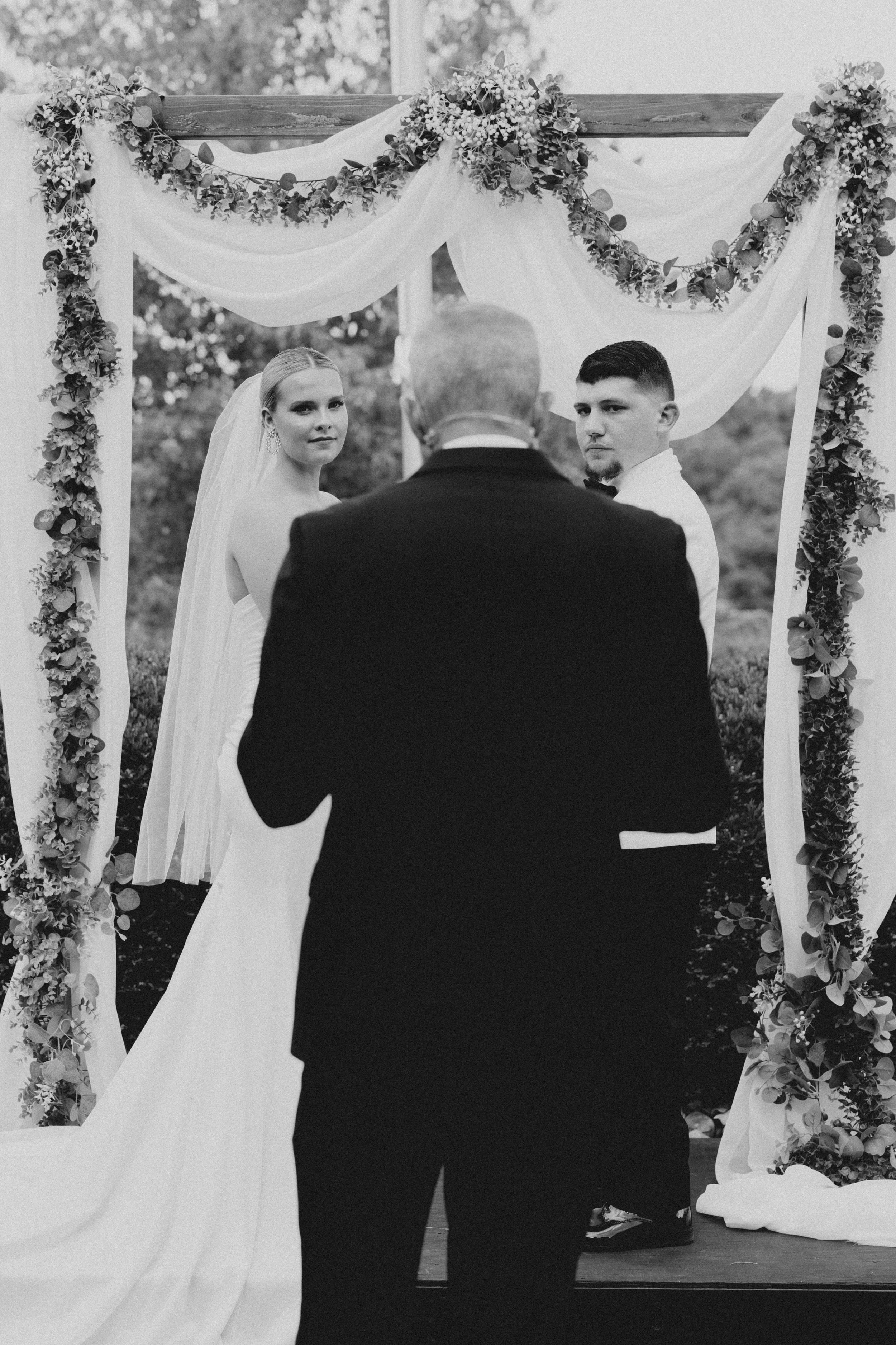 A black and white photo of a wedding ceremony showing a bride and groom facing an officiant, with floral and fabric decorations hanging on a wooden arch behind them.
