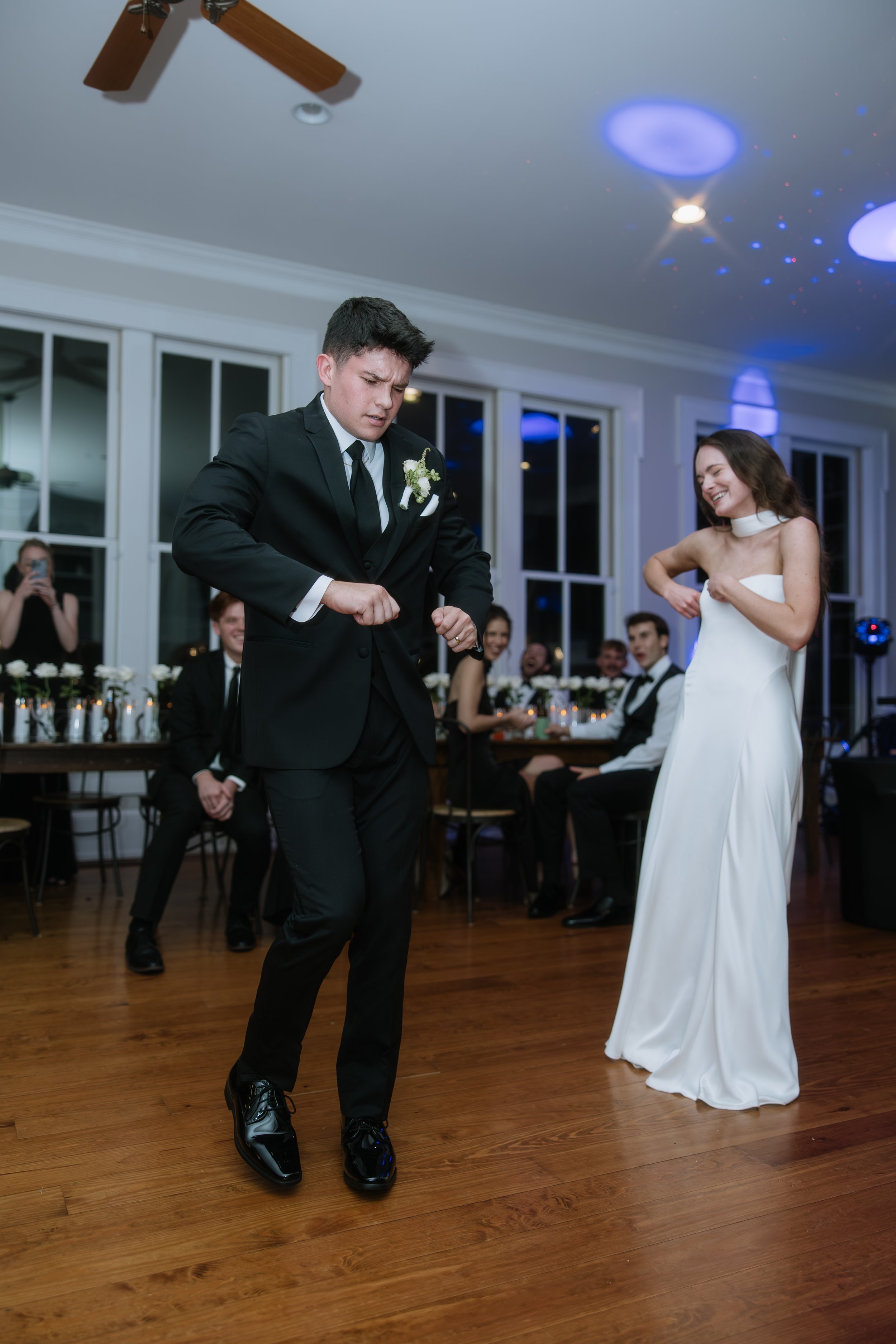 A groom in a black tuxedo dancing with a woman in a white wedding dress at a wedding reception. Guests are seated at tables in the background, and a woman is taking a photo. The room has white walls, large windows, and a wooden floor.