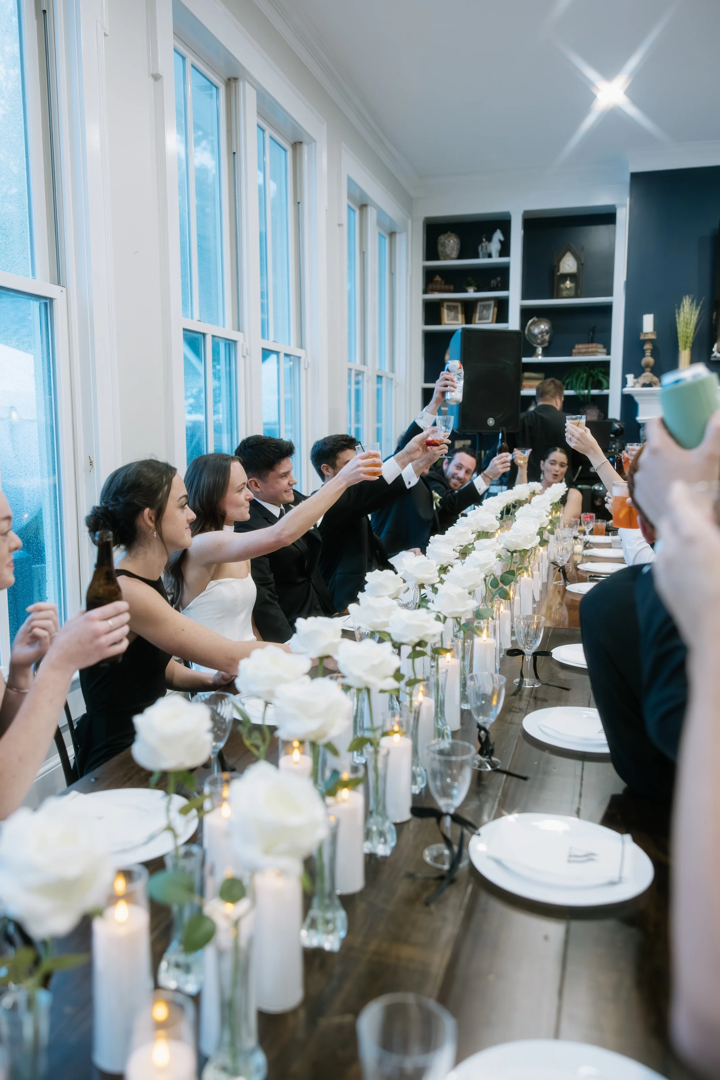 People at a wedding reception raising their glasses for a toast, seated at a long table decorated with white roses and candles.