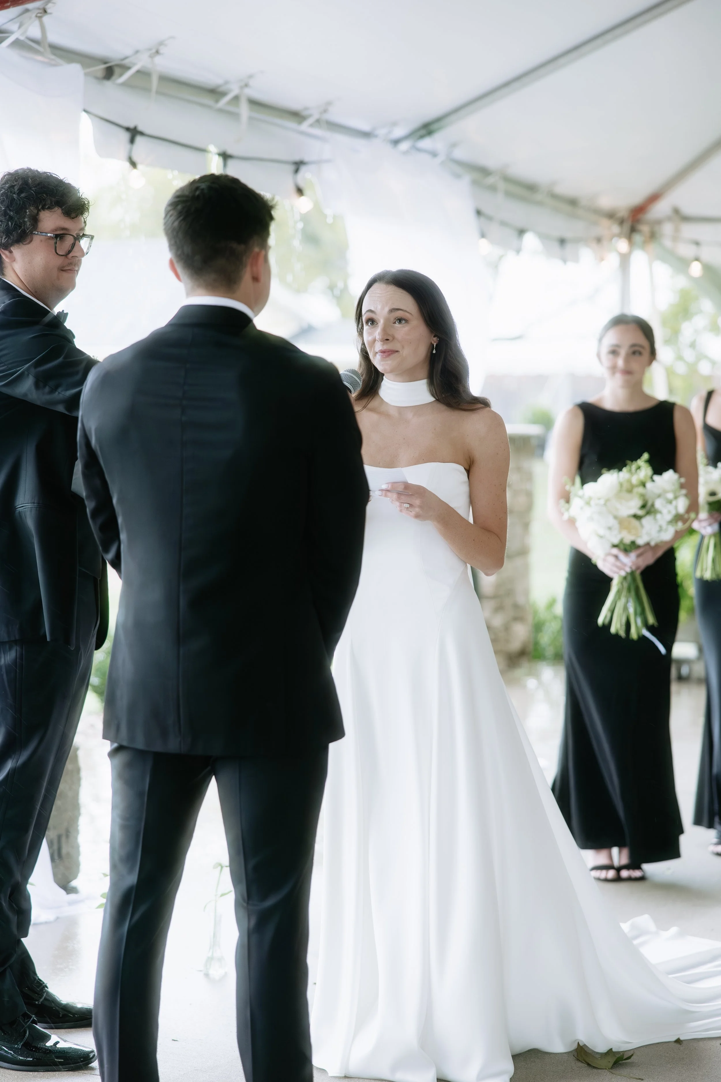 A bride in a white wedding dress and a woman in a black dress holding a bouquet, standing under a tent, during a wedding ceremony.